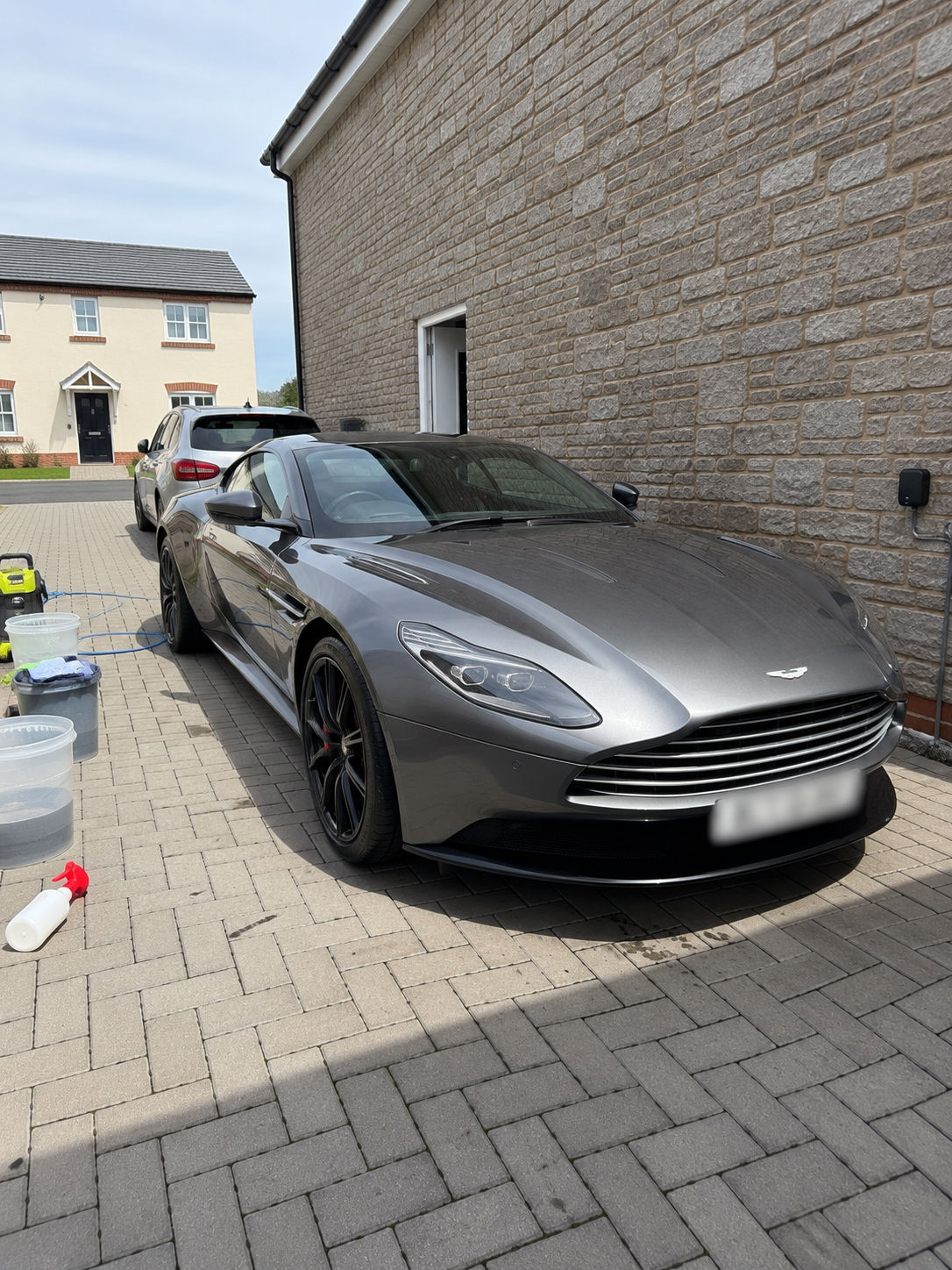 A gray Aston Martin sports car parked on a driveway beside a brick house, with car washing supplies and buckets nearby.