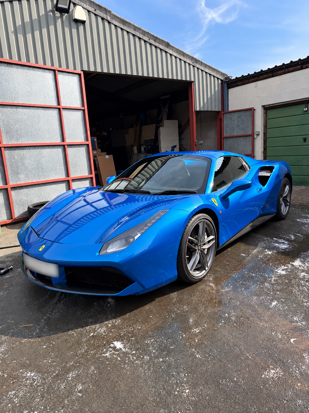 Blue Ferrari sports car parked outdoors near a garage with a open door and green door, under a blue sky with wispy clouds.