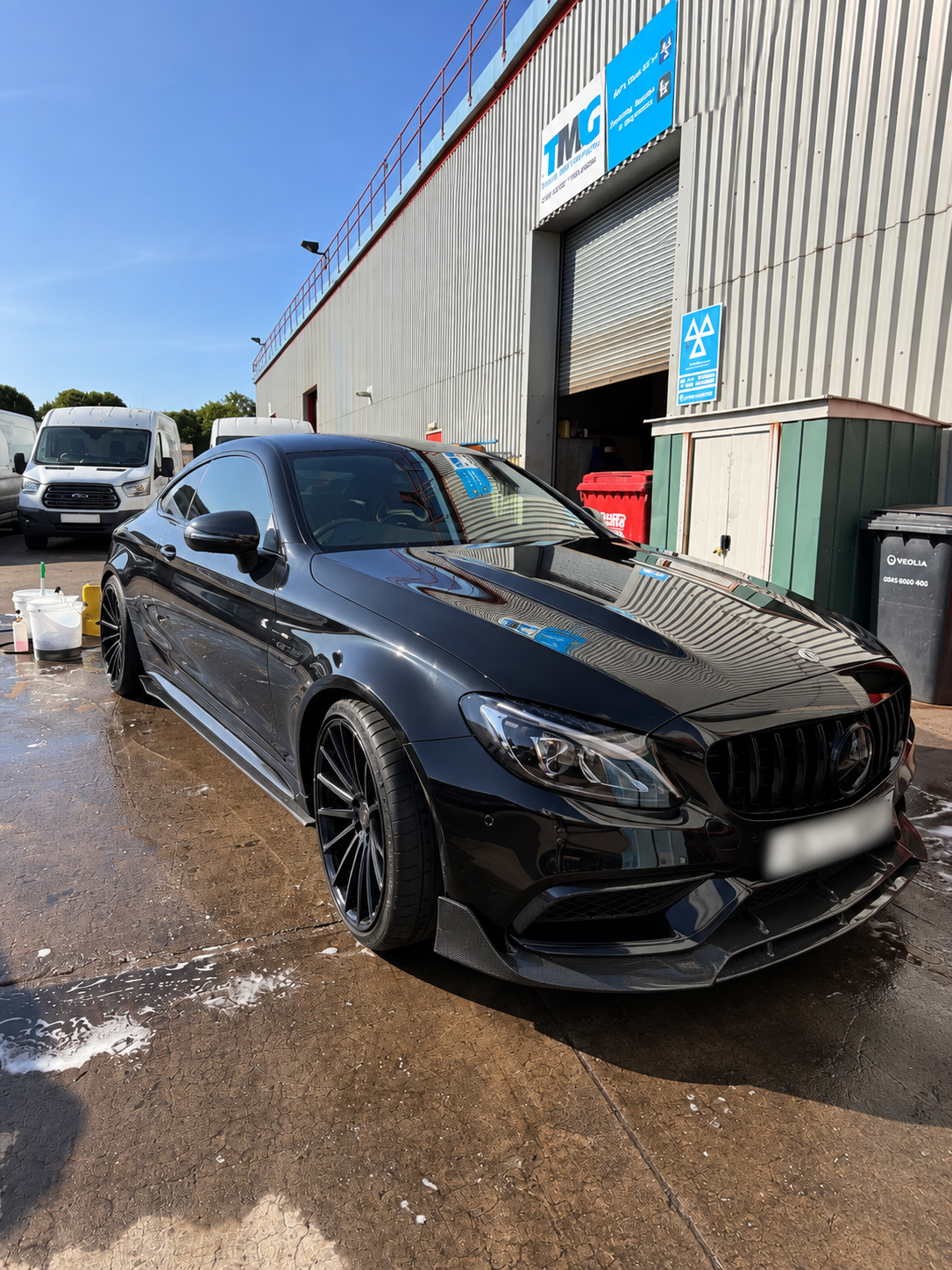 Black sports car at an auto wash station with soap suds on the ground, building with a closed roller door, and other vehicles in the background.