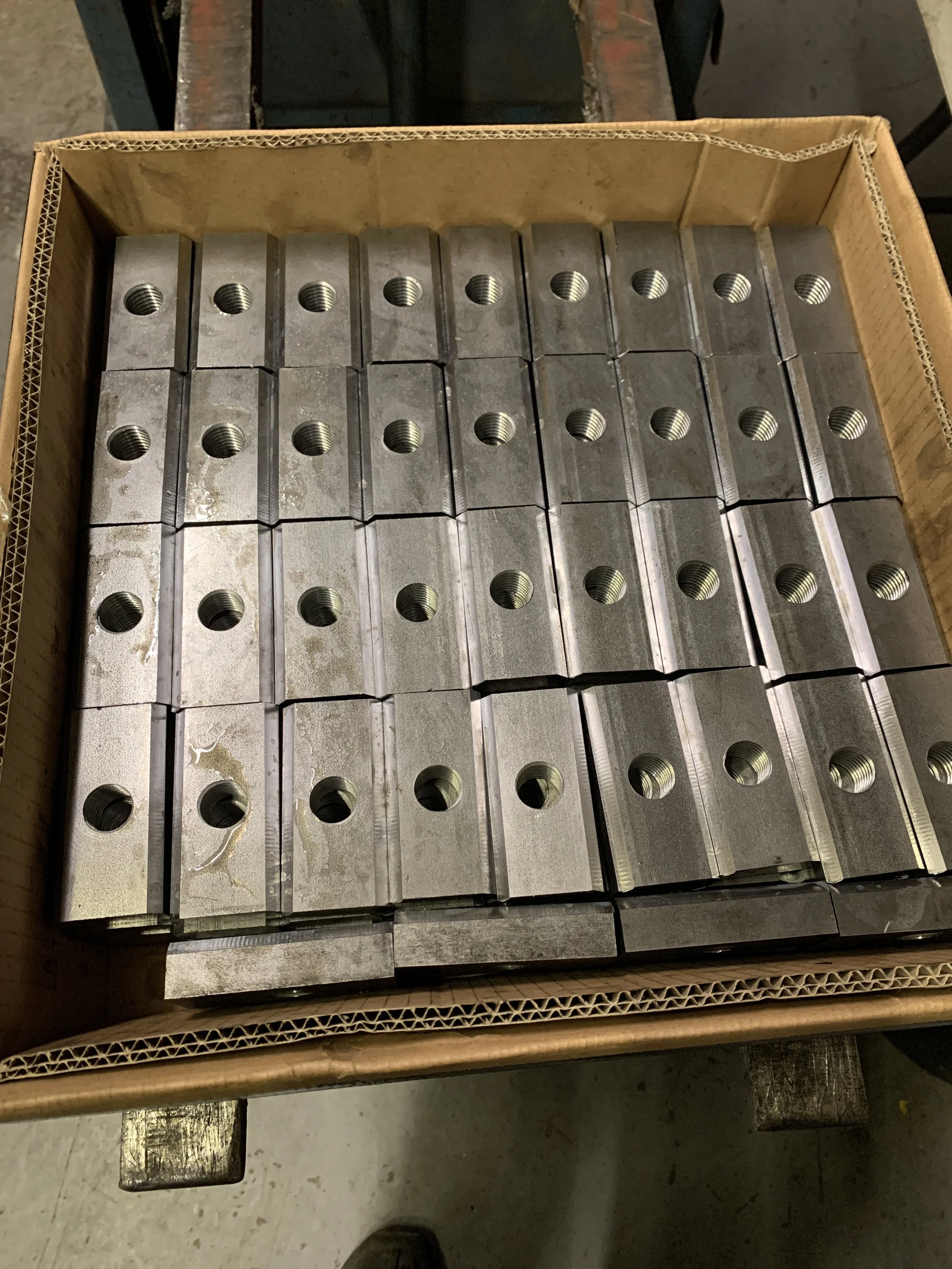 A cardboard box filled with multiple metallic metal parts with threaded holes.
