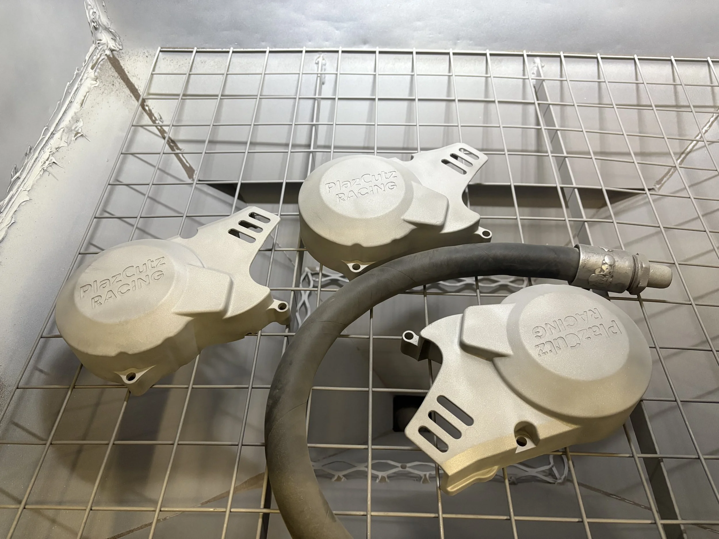 Three white Plazmuz Racing brake calipers and one used black brake hose on a metal wire grid surface.