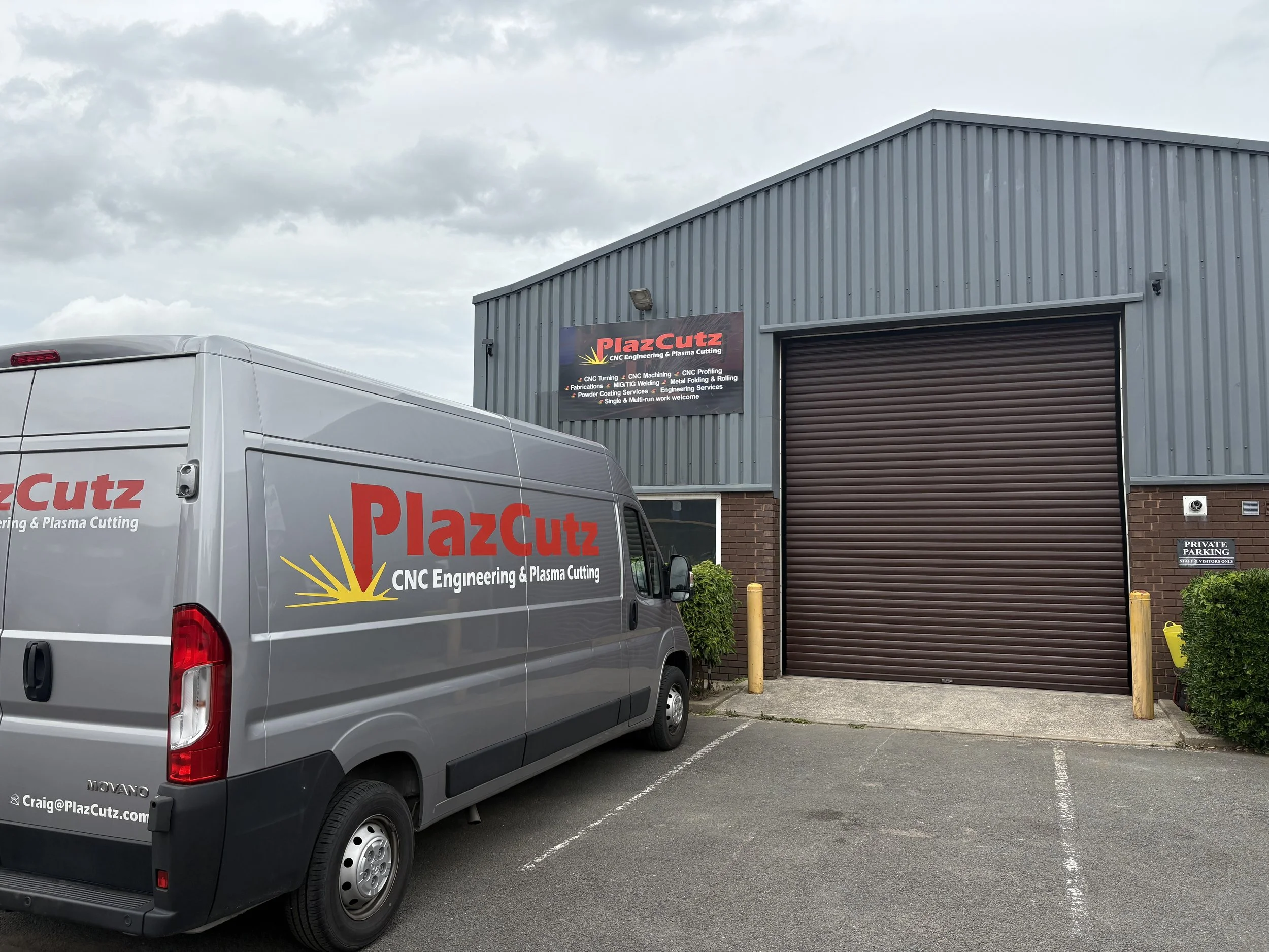 Photo of a gray PlazCutz service van parked in front of a metal industrial building with a large roll-up door. The van has the company's logo and contact information. The building has a sign showing the company's services, and there's a sign indicating private parking.