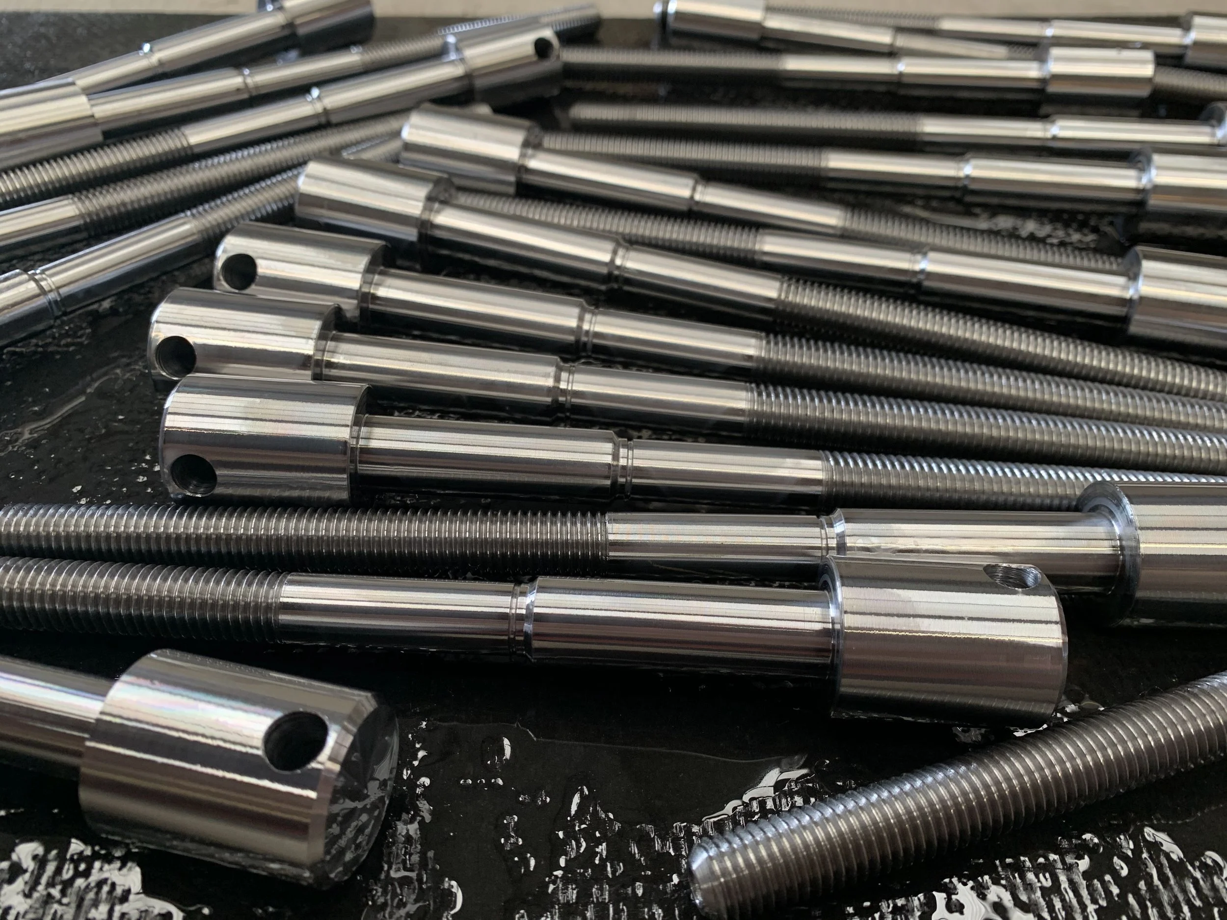 Close-up of multiple shiny, cylindrical metal rods with threaded sections and holes, laid out on a dark surface.