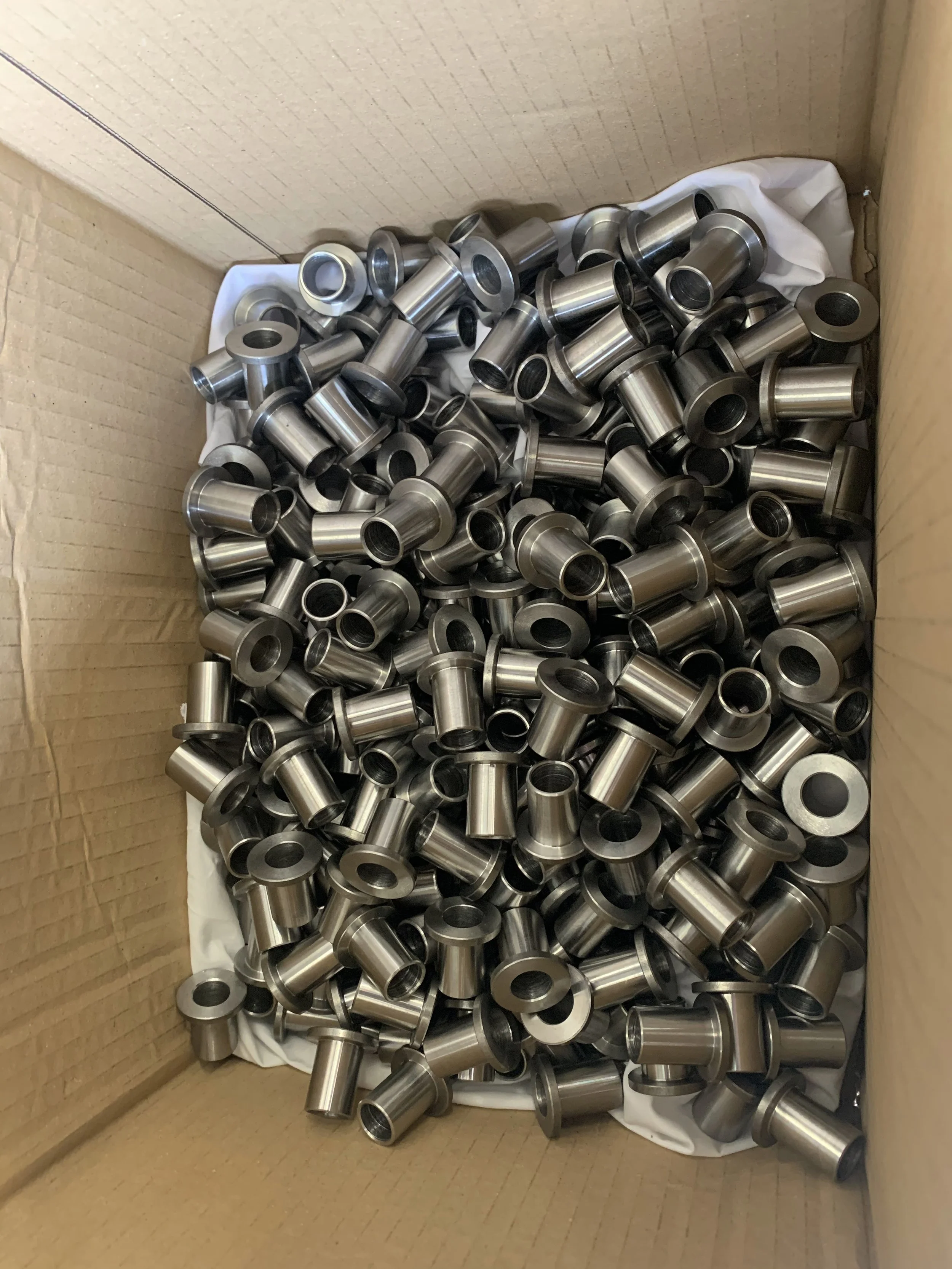 A cardboard box filled with small, shiny, metallic cylindrical components, likely machine parts or hardware, stacked on a white inner lining.
