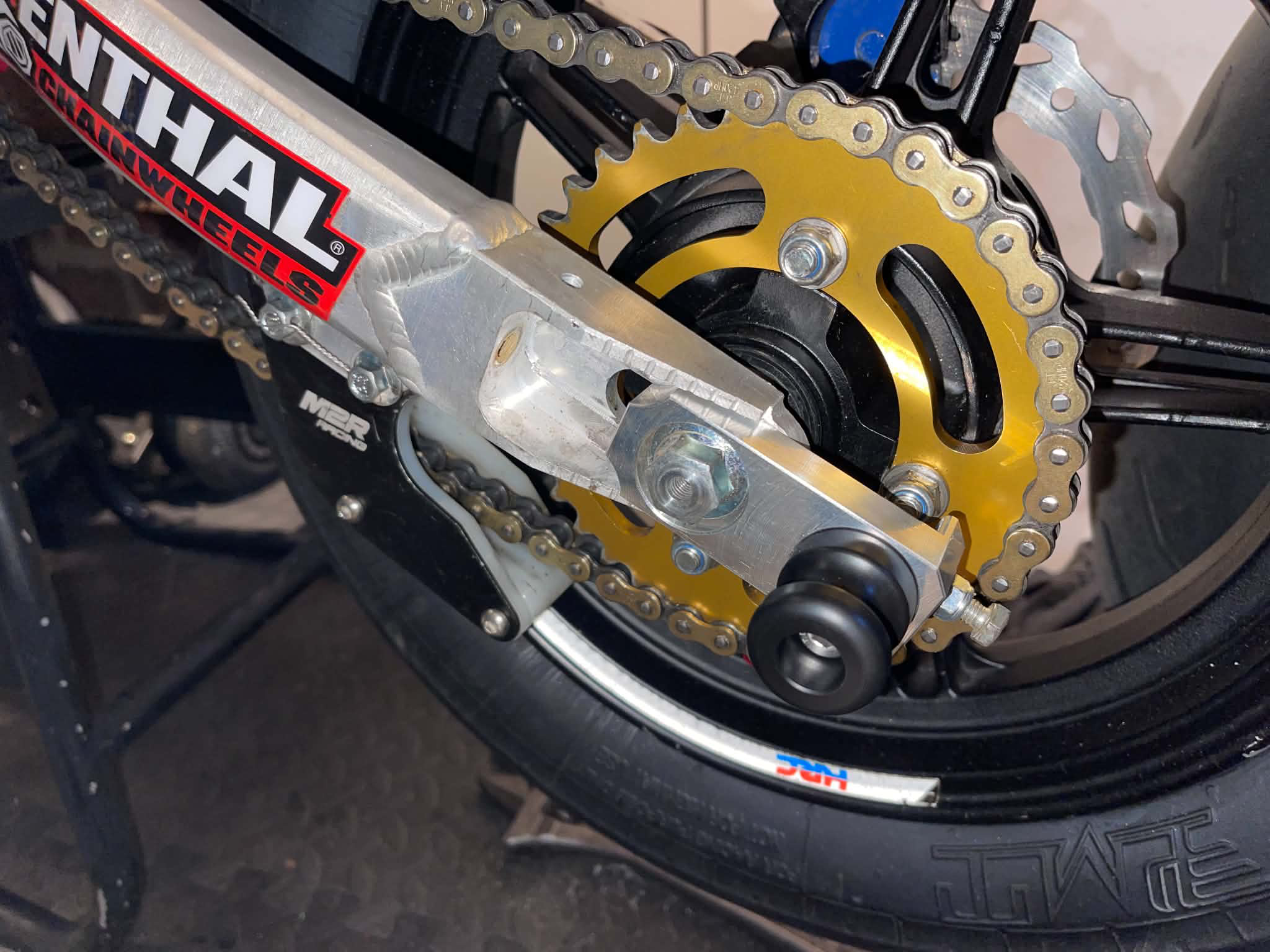 Rear chain adjuster block with lifting bobbin fitted to a pitbike wheel, improving chain adjustment and paddock stand use.