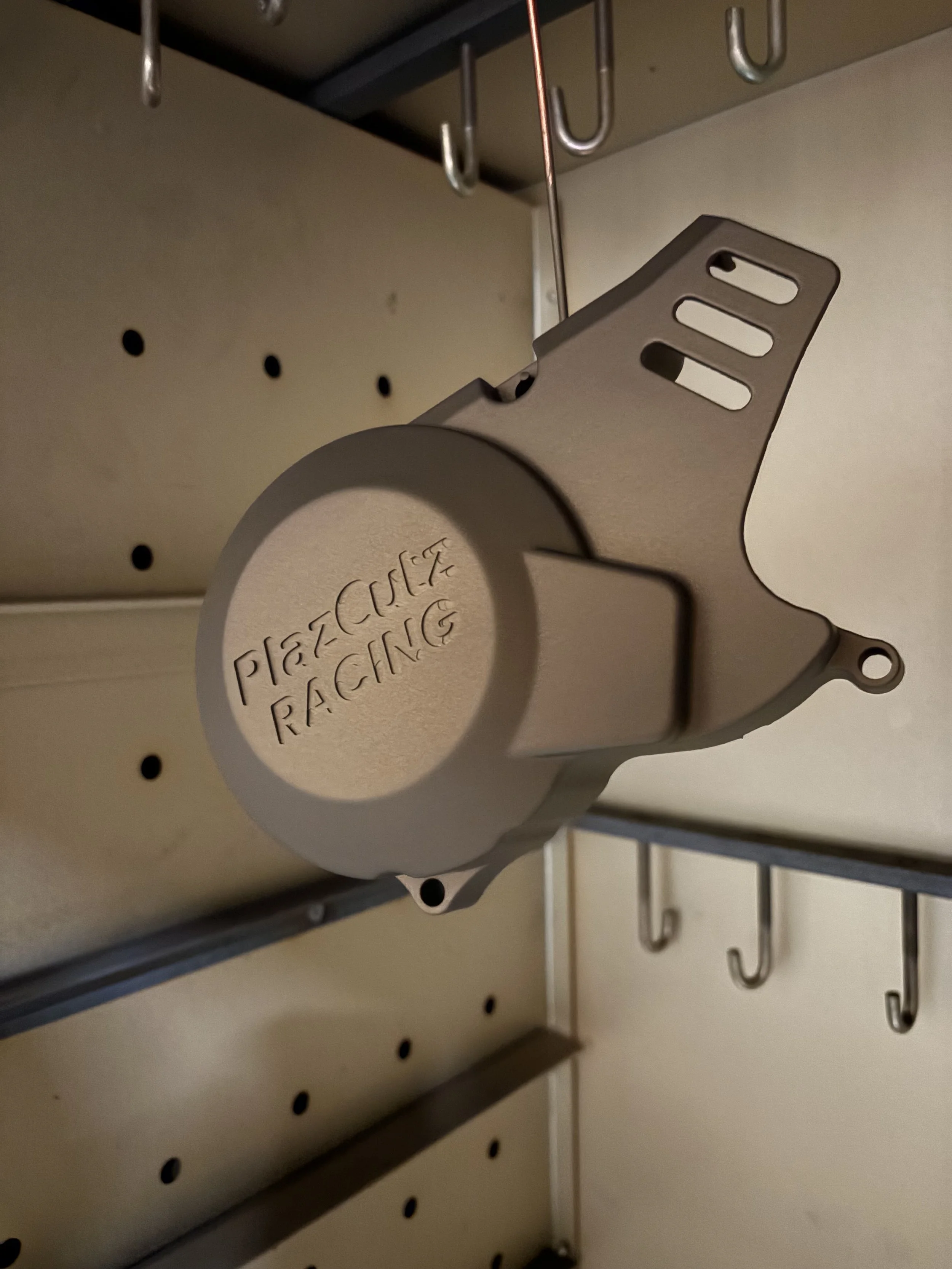 A beige, circular brake caliper part with the engraved text 'PLAZCUTZ RACING' hanging on a peg in a tool storage area.