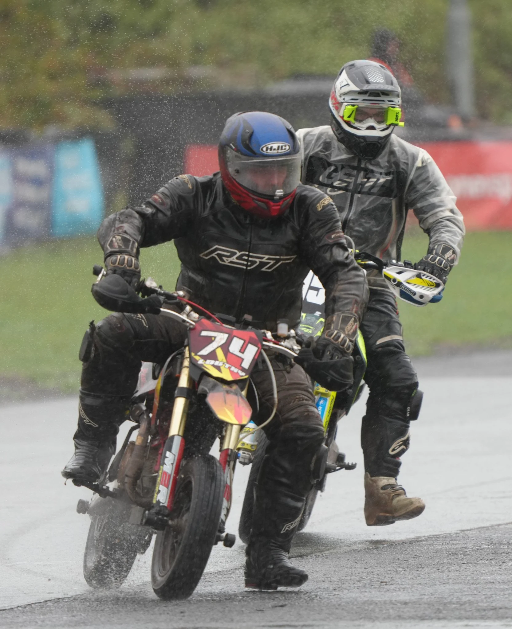 Two motorcyclists riding in the rain, one in the front on a dirt bike with the number 74, and a second rider behind, both wearing protective gear.