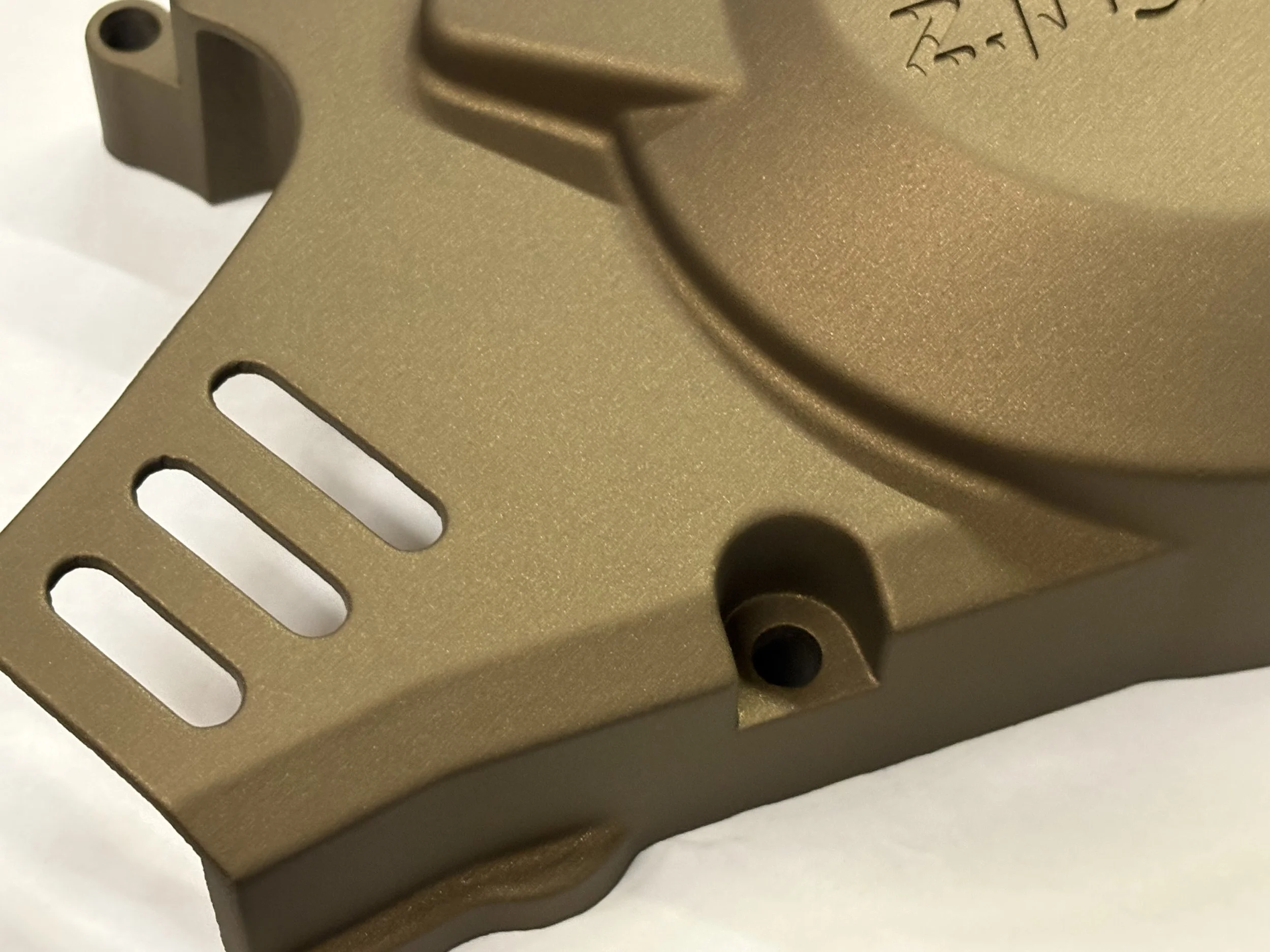 Close-up of a metallic, tan-colored object with elongated slots and screw holes, possibly a component of a mechanical or electronic device.