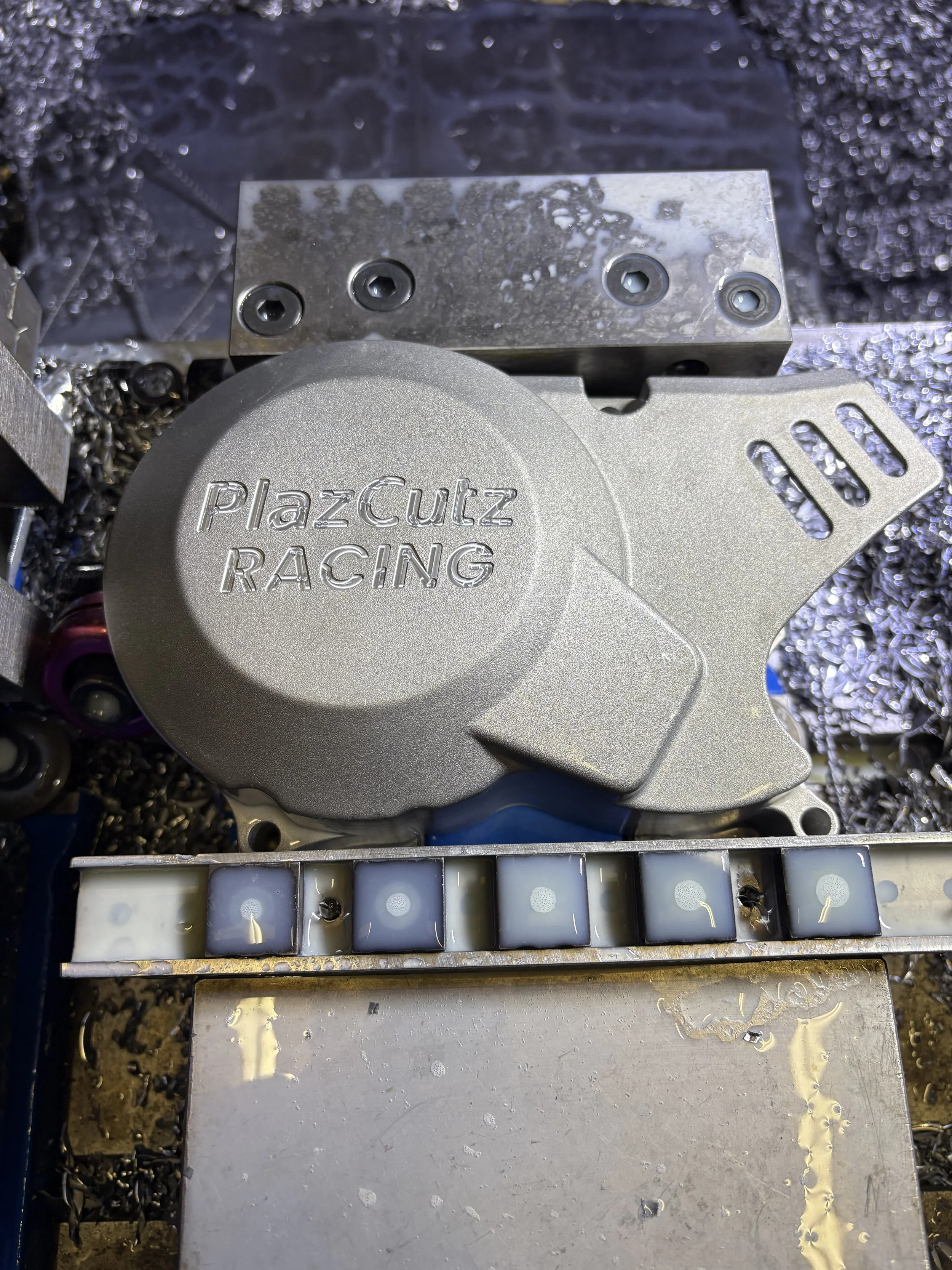 Close-up of a PlazCutz Racing motor or engine part with metal components and a row of small electronic ignition modules.