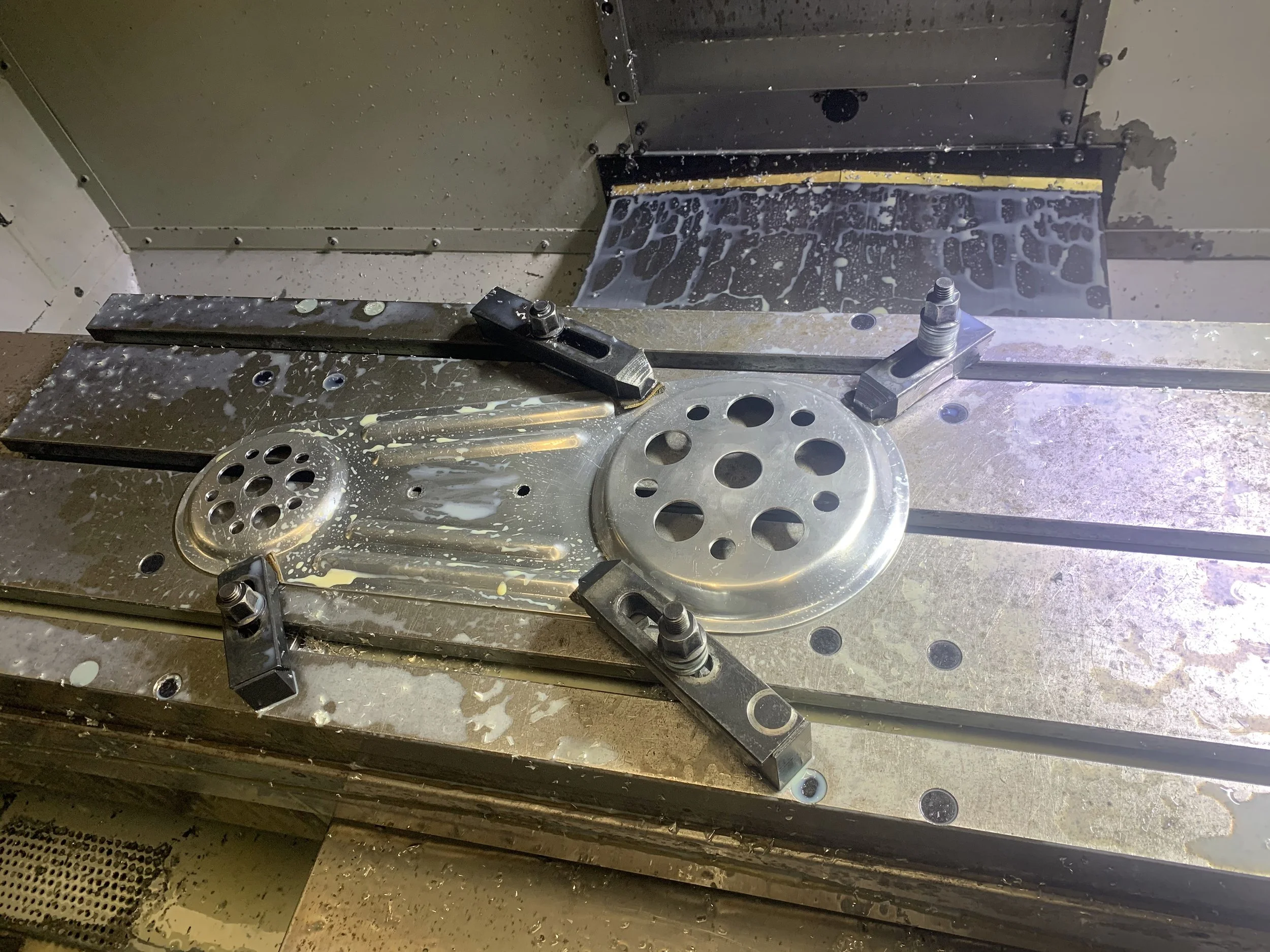 Metal machining setup with metal parts, clamps, and a milling machine table covered in coolant.