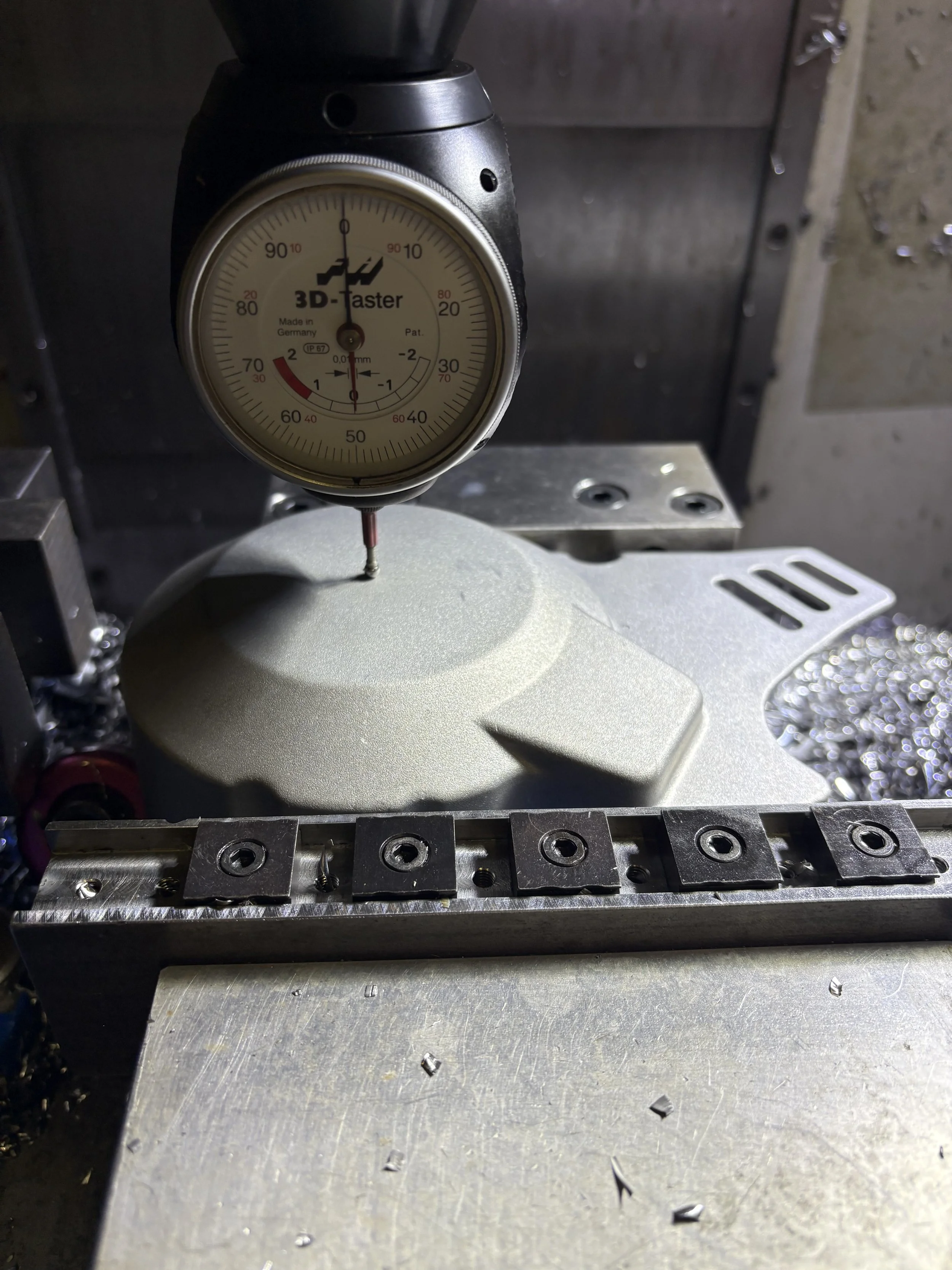 A precision dial gauge measuring the flatness of a machined metal surface. The gauge pin contacts the surface, while the dial shows readings in millimeters.