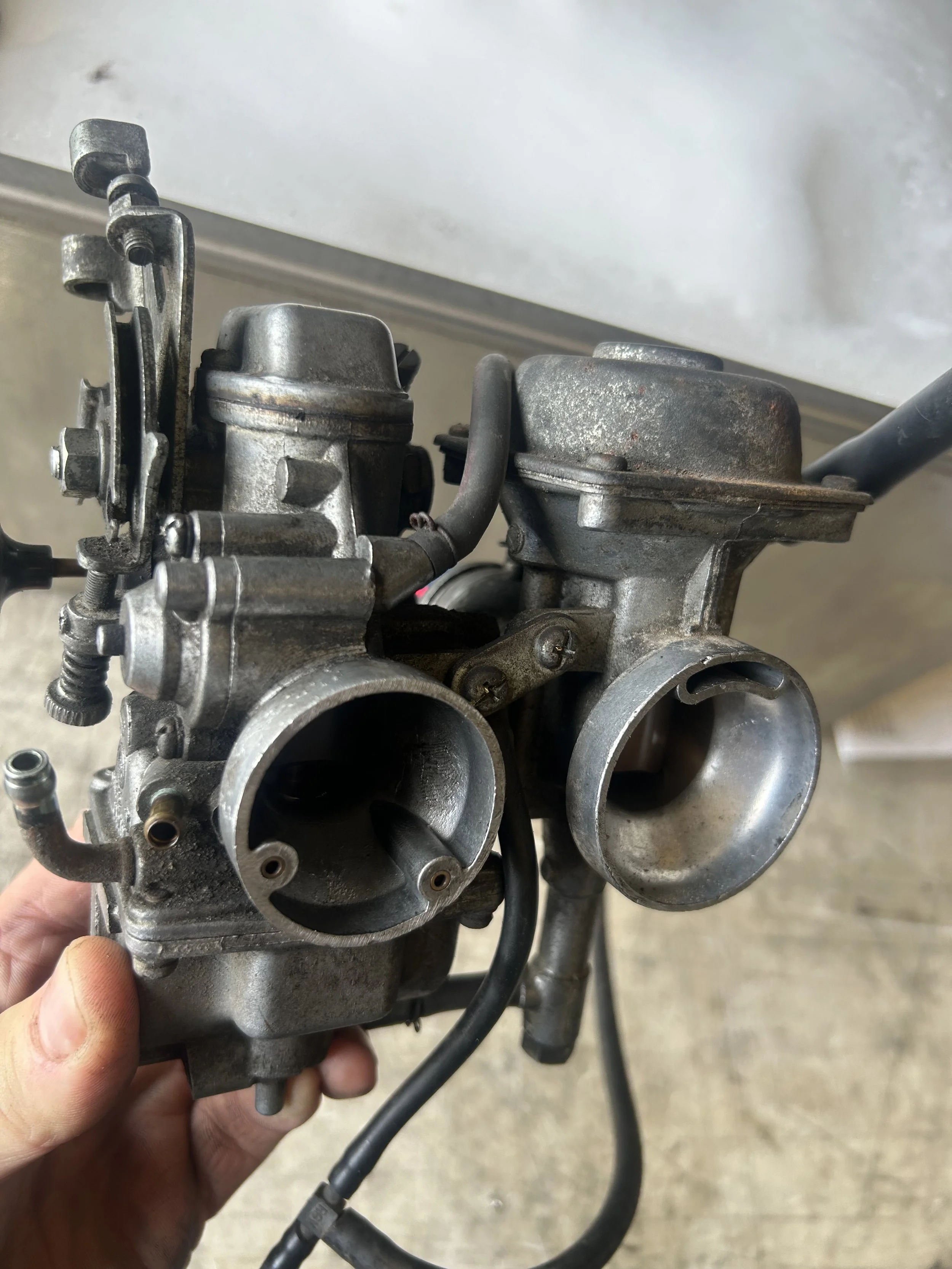 A pair of vintage motorcycle carburetors held in hand, showing metal construction and openings for air or fuel intake.