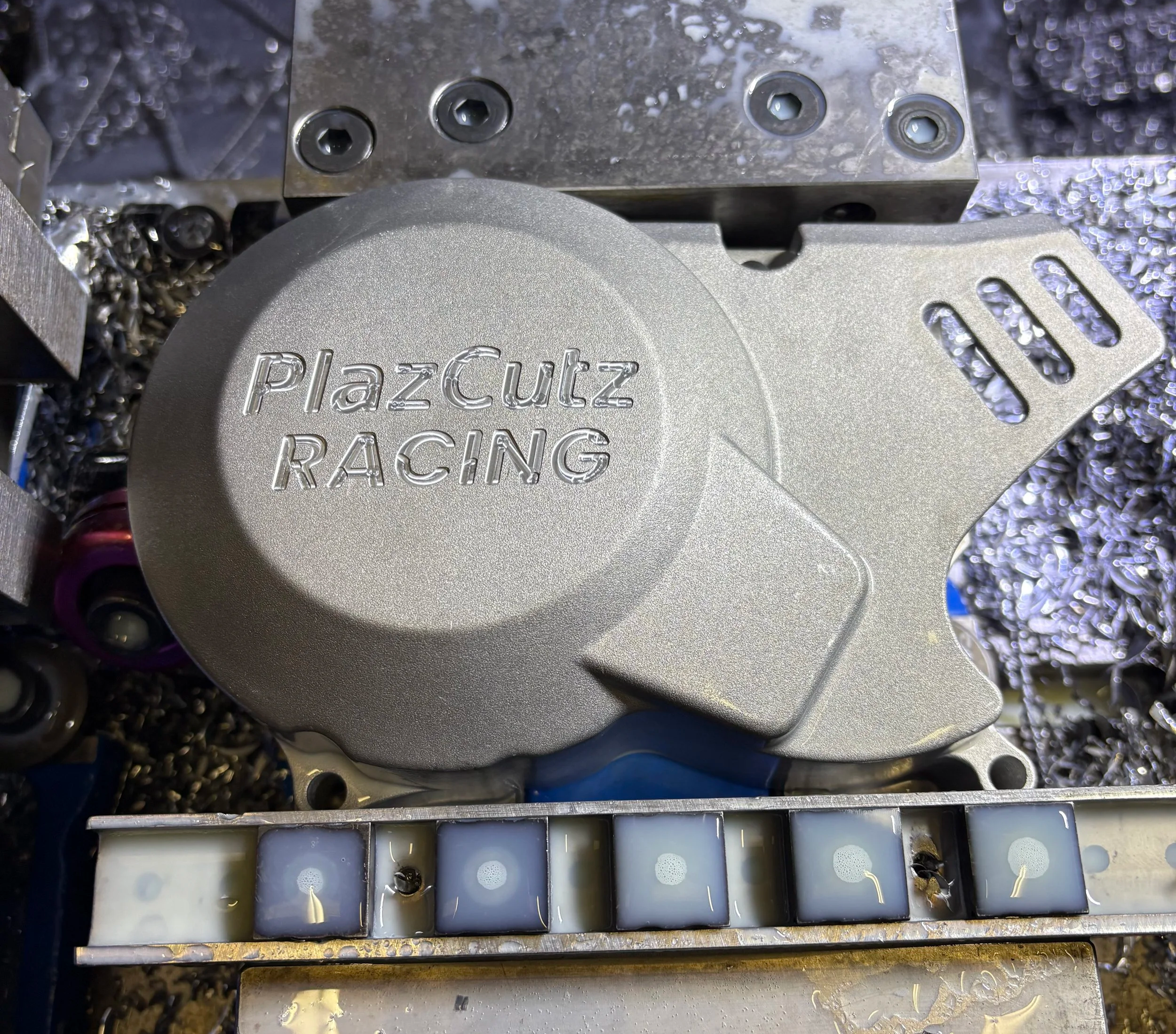 Close-up of a metallic engine part labeled 'PlazCutz RACING' with various screws and components around it.