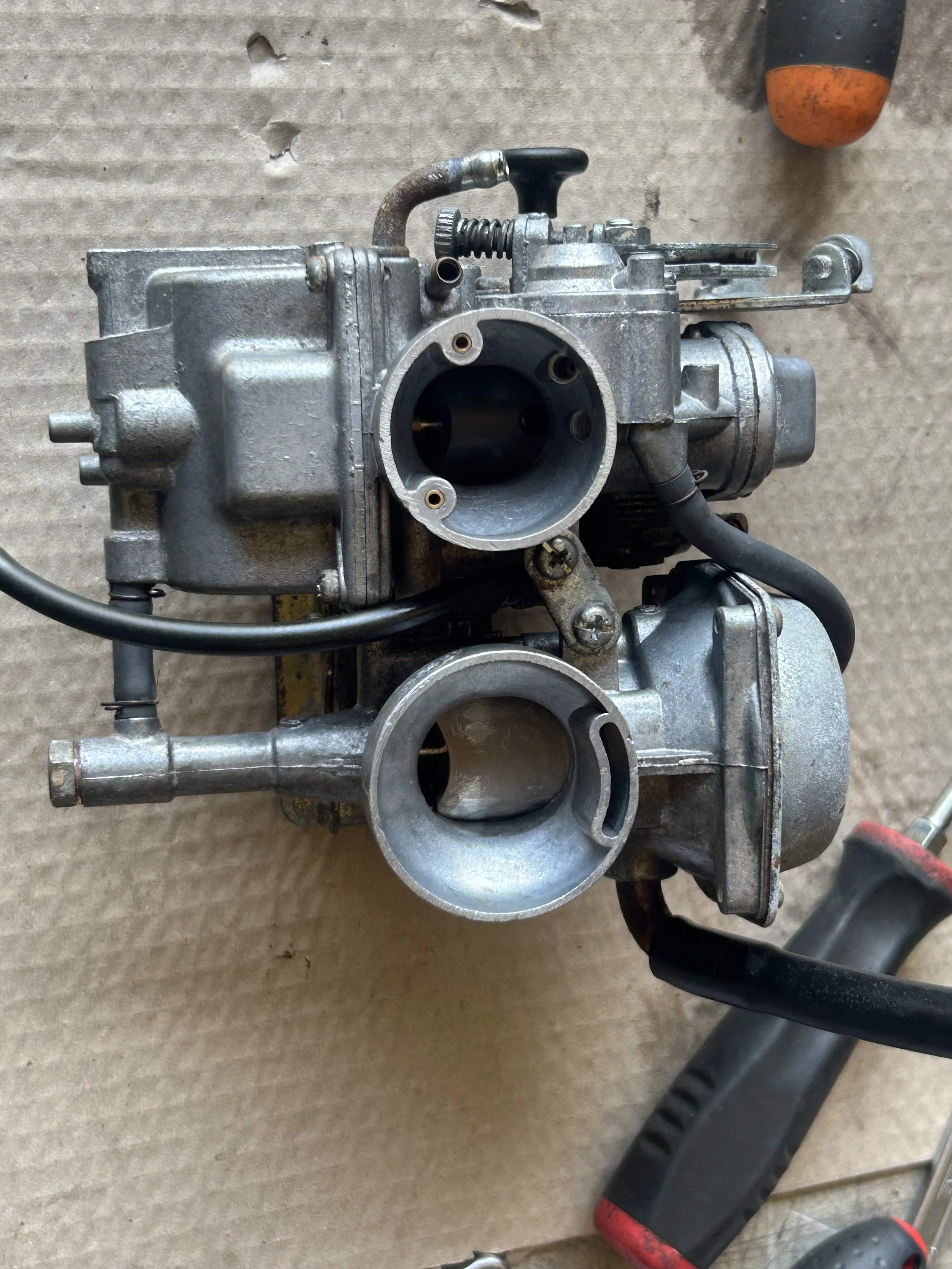 A disassembled automotive carburetor placed on a cardboard surface next to a screwdriver.