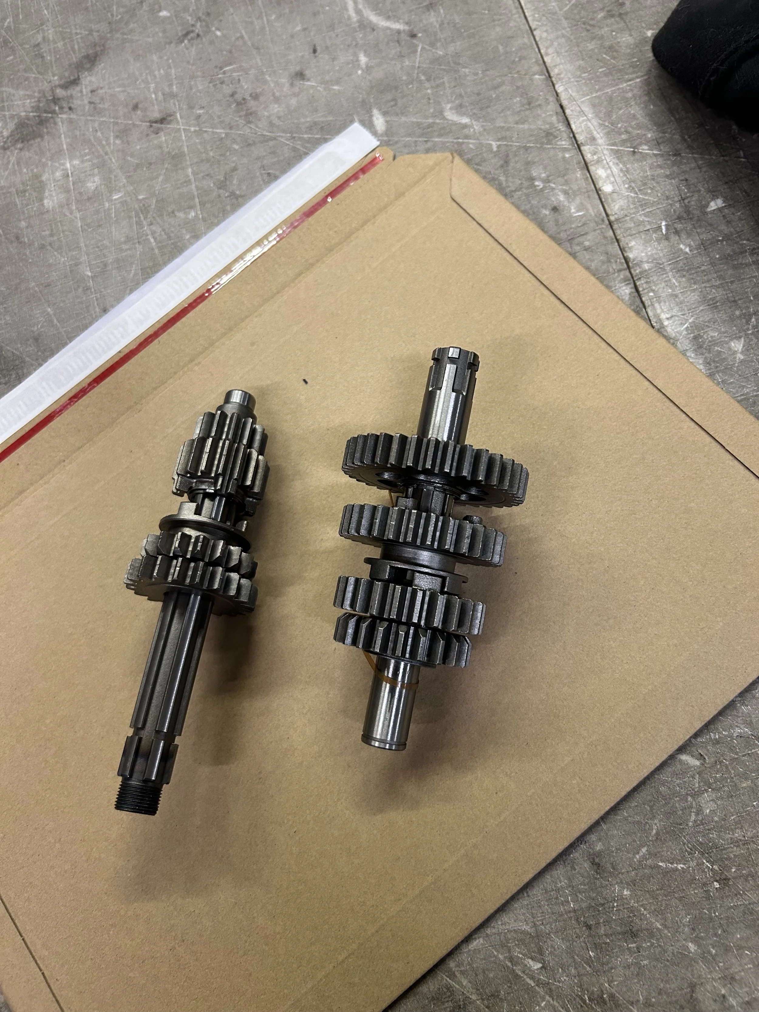 Two metal gear shafts with multiple gears mounted on them, placed on a piece of cardboard on a metallic surface.