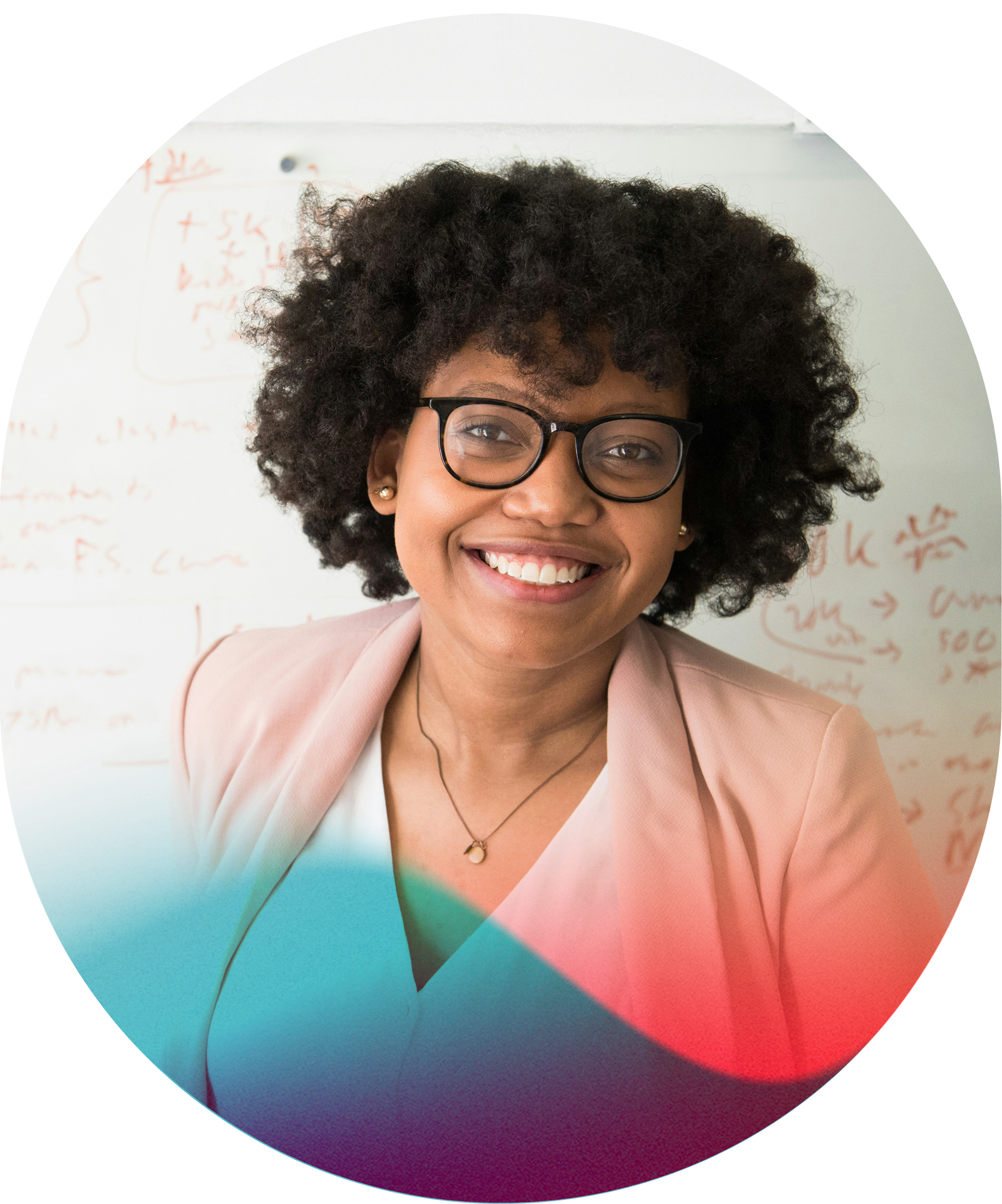 A smiling woman with curly black hair and glasses, wearing a light blazer, standing in front of a whiteboard with red handwriting.