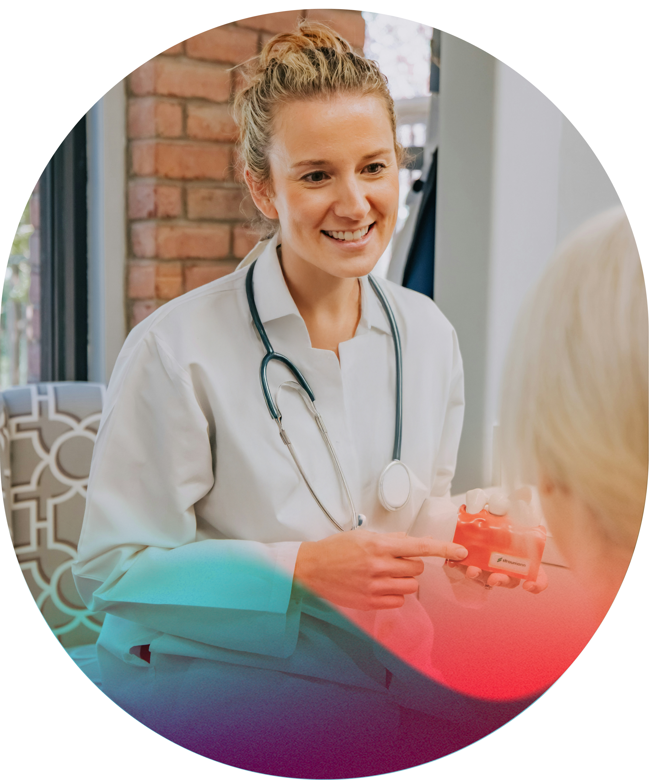 A female doctor with blonde hair tied back, wearing a white coat and stethoscope, smiling and talking to an elderly woman with blonde hair in a medical office or clinic.