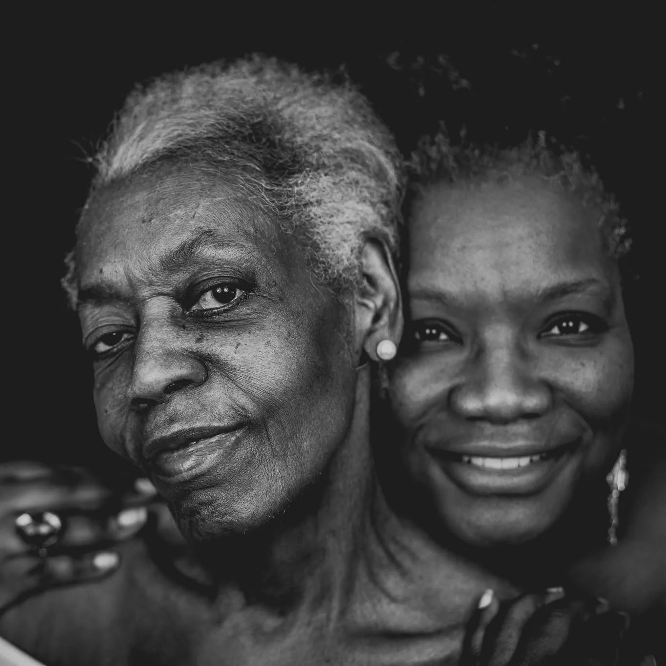 Close-up black and white photo of an elderly woman with gray hair and a young girl with curly hair smiling, both facing the camera.