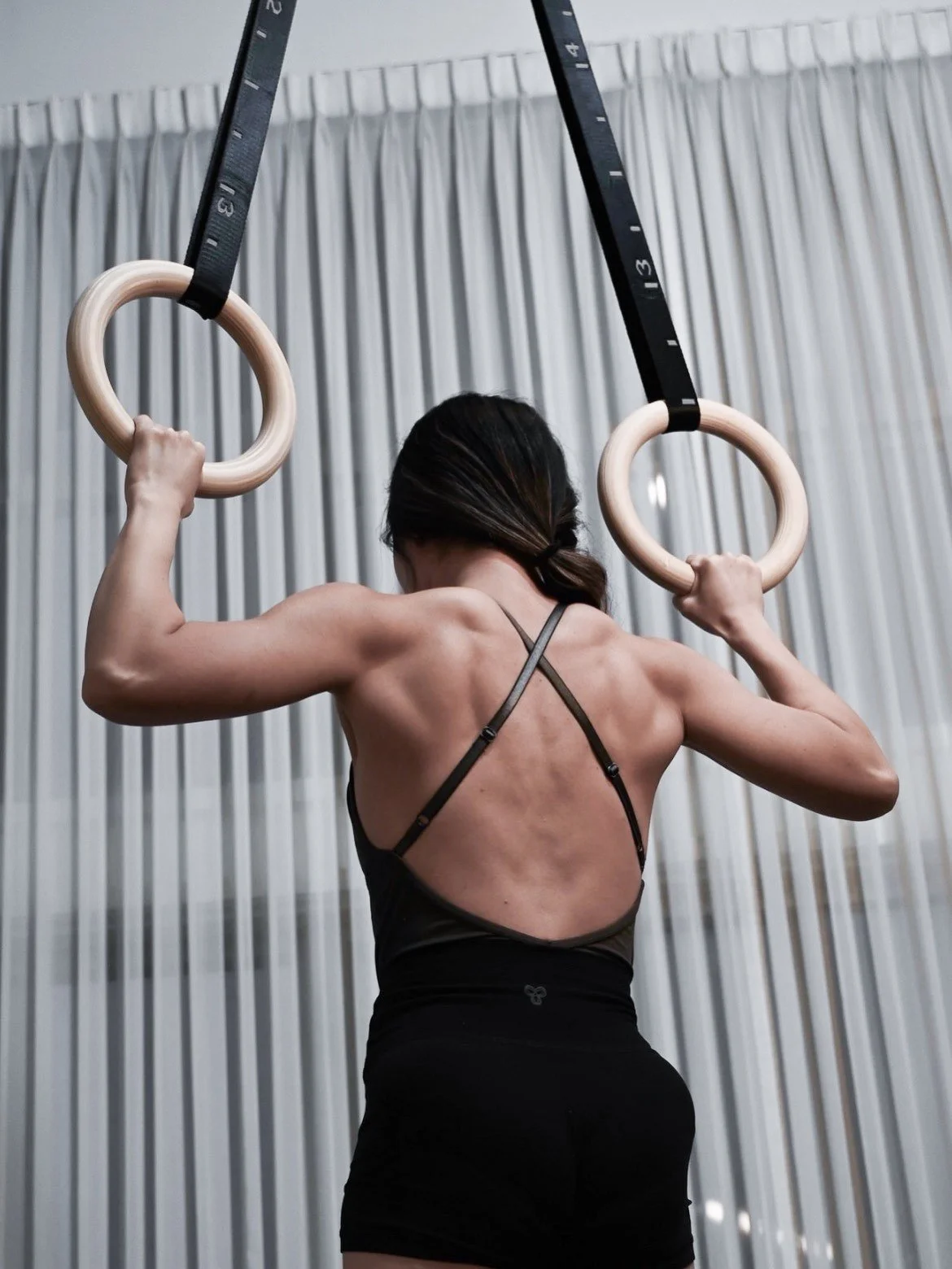 A woman exercising pull up on gymnastic rings, seen from the back, wearing a black sports bra and black shorts, with a grey curtain in the background.