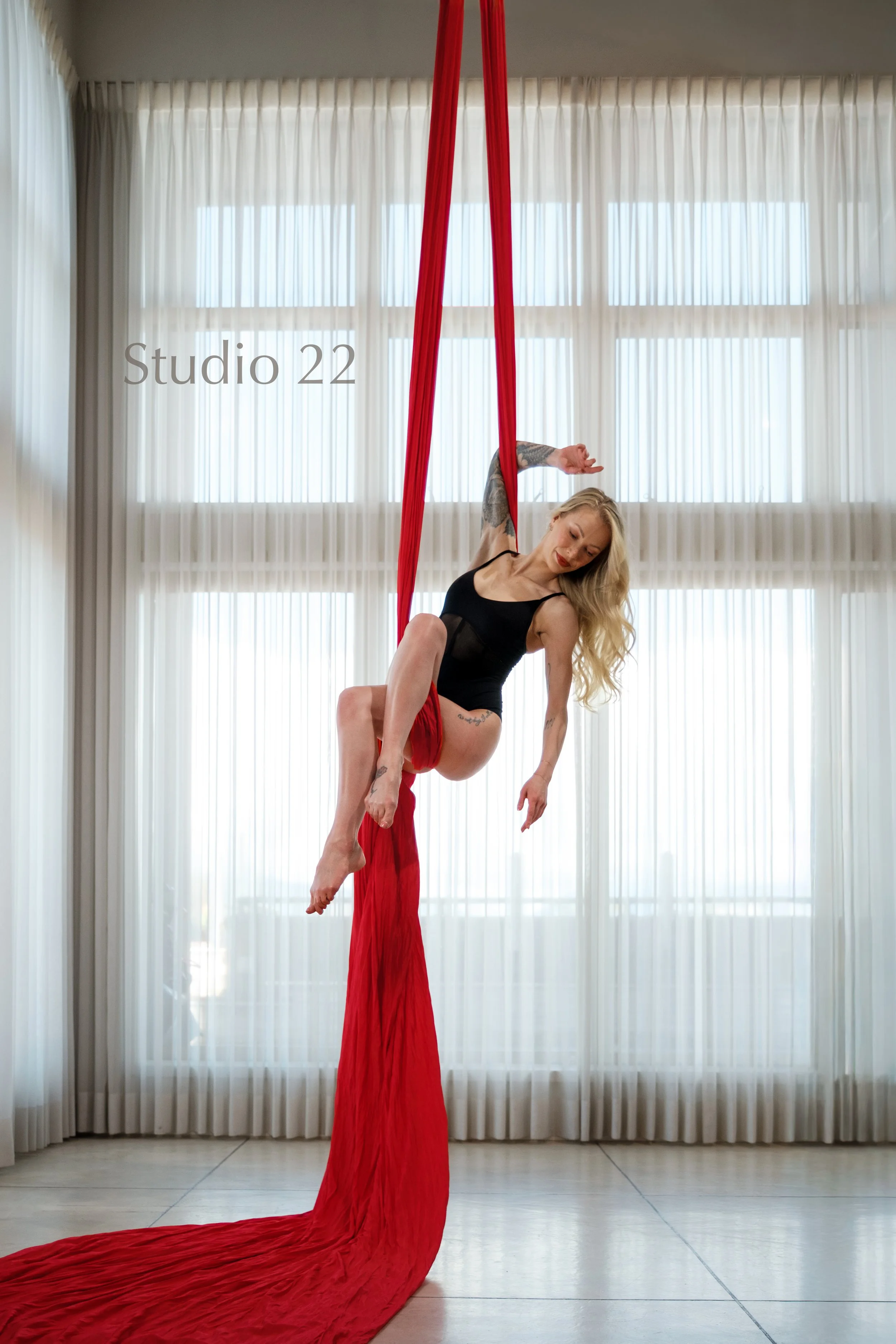 A woman performing aerial silks acrobatics in a studio with large windows and sheer curtains in Vancouver.