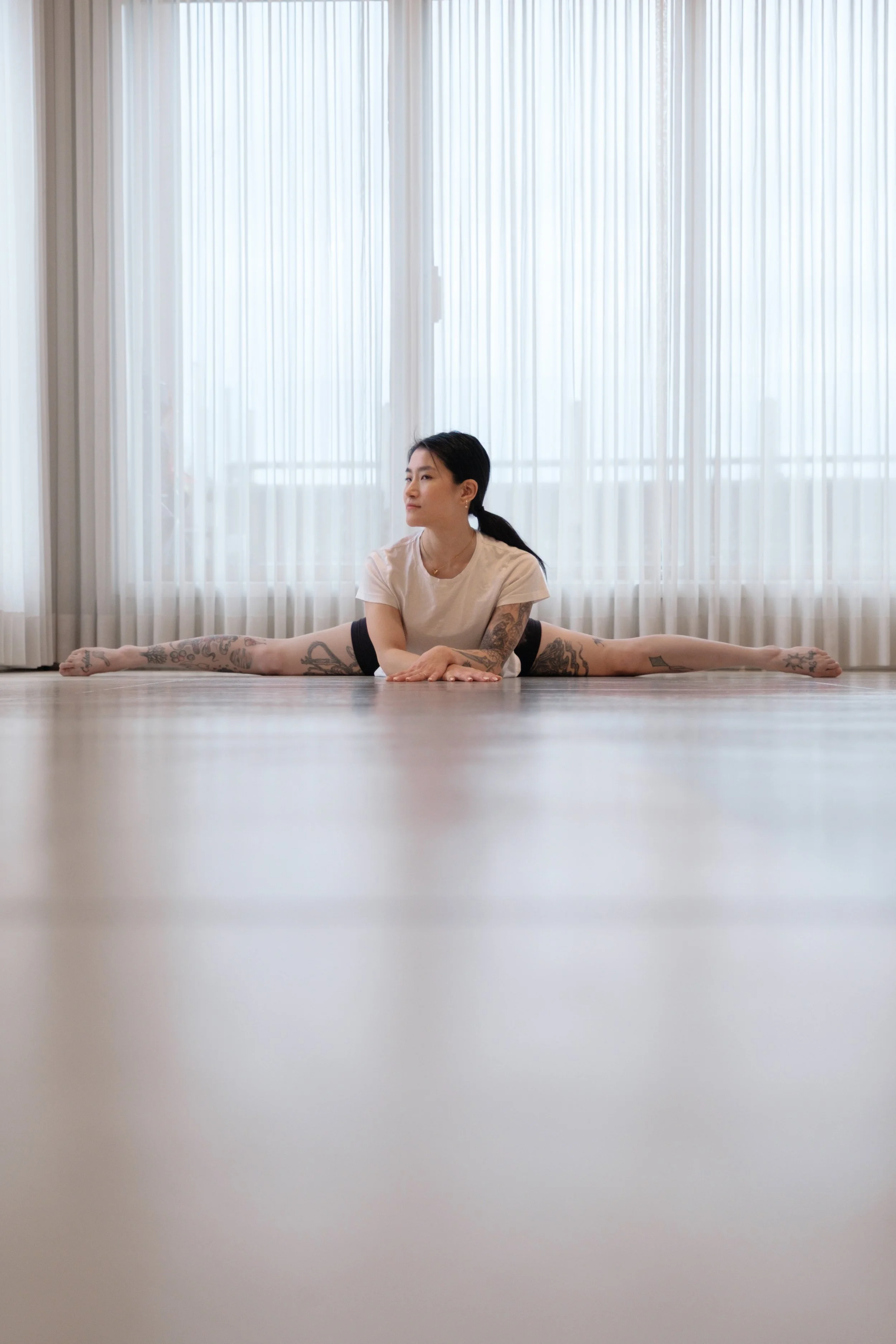 A woman doing a middle split yoga pose on a wooden floor in a bright room with large windows and sheer curtains.