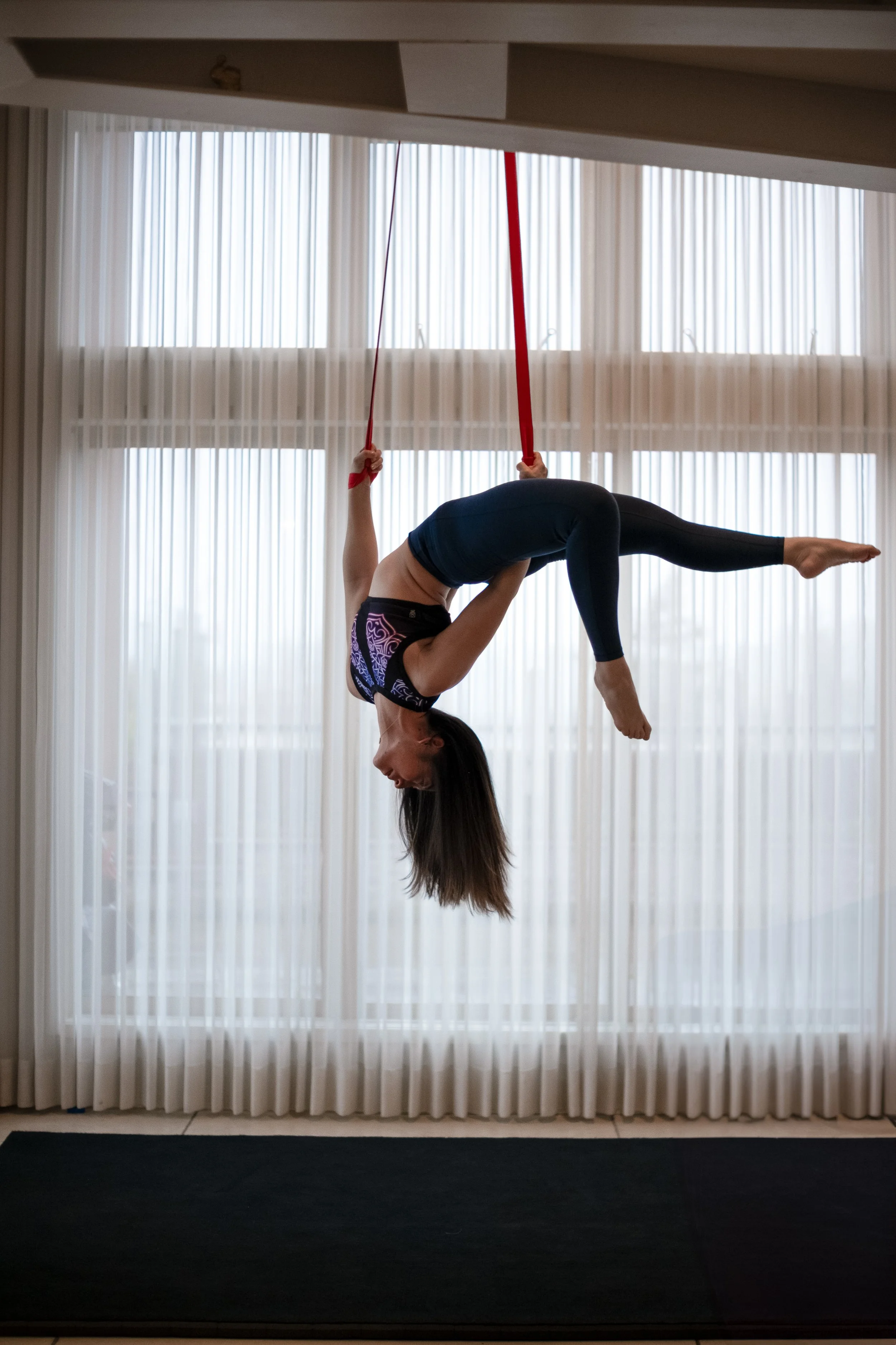 A woman performing an aerial straps pose upside down in front of a large window with sheer white curtains.