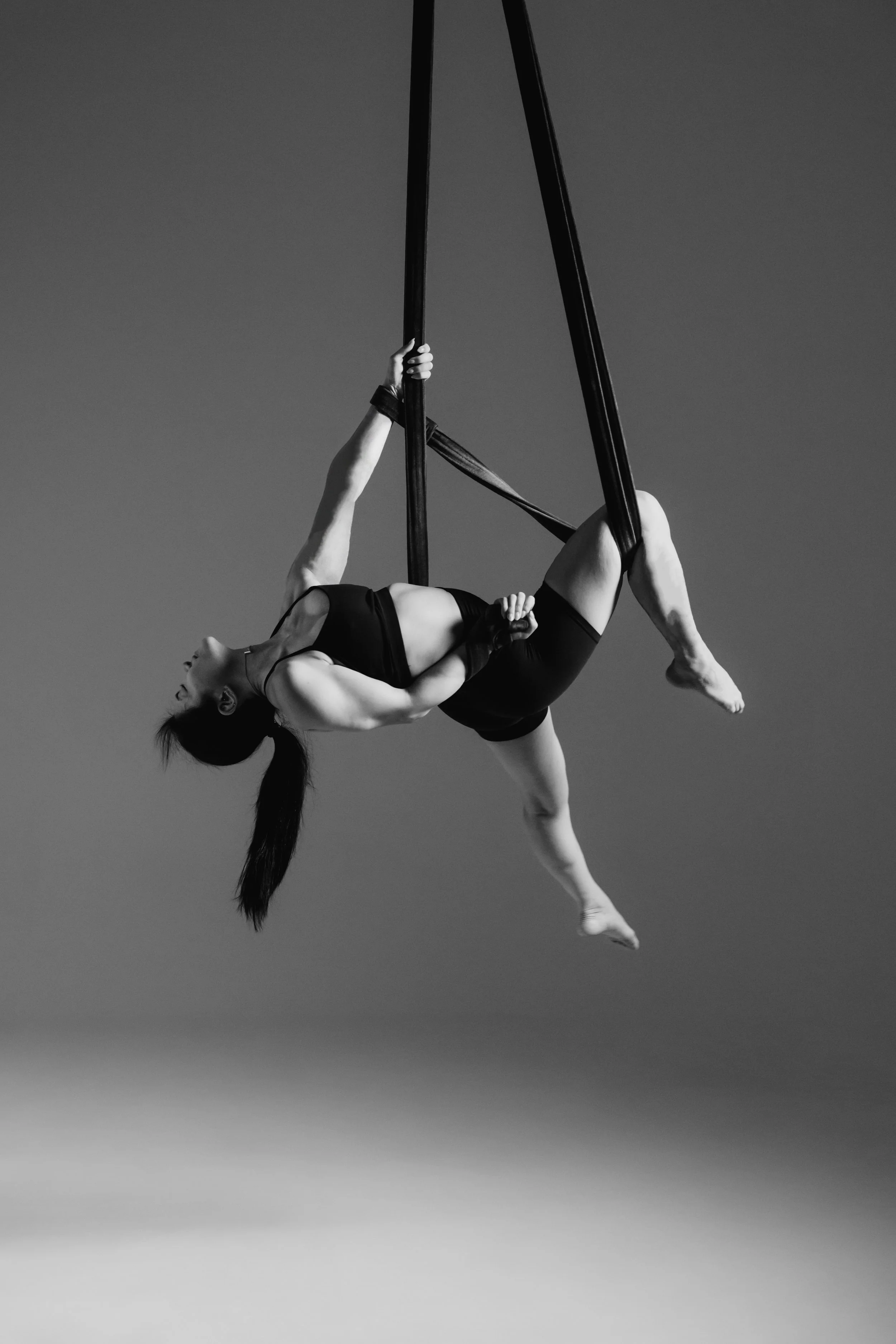 Black and white photo of a female aerialist performing splits on aerial straps against a plain background.