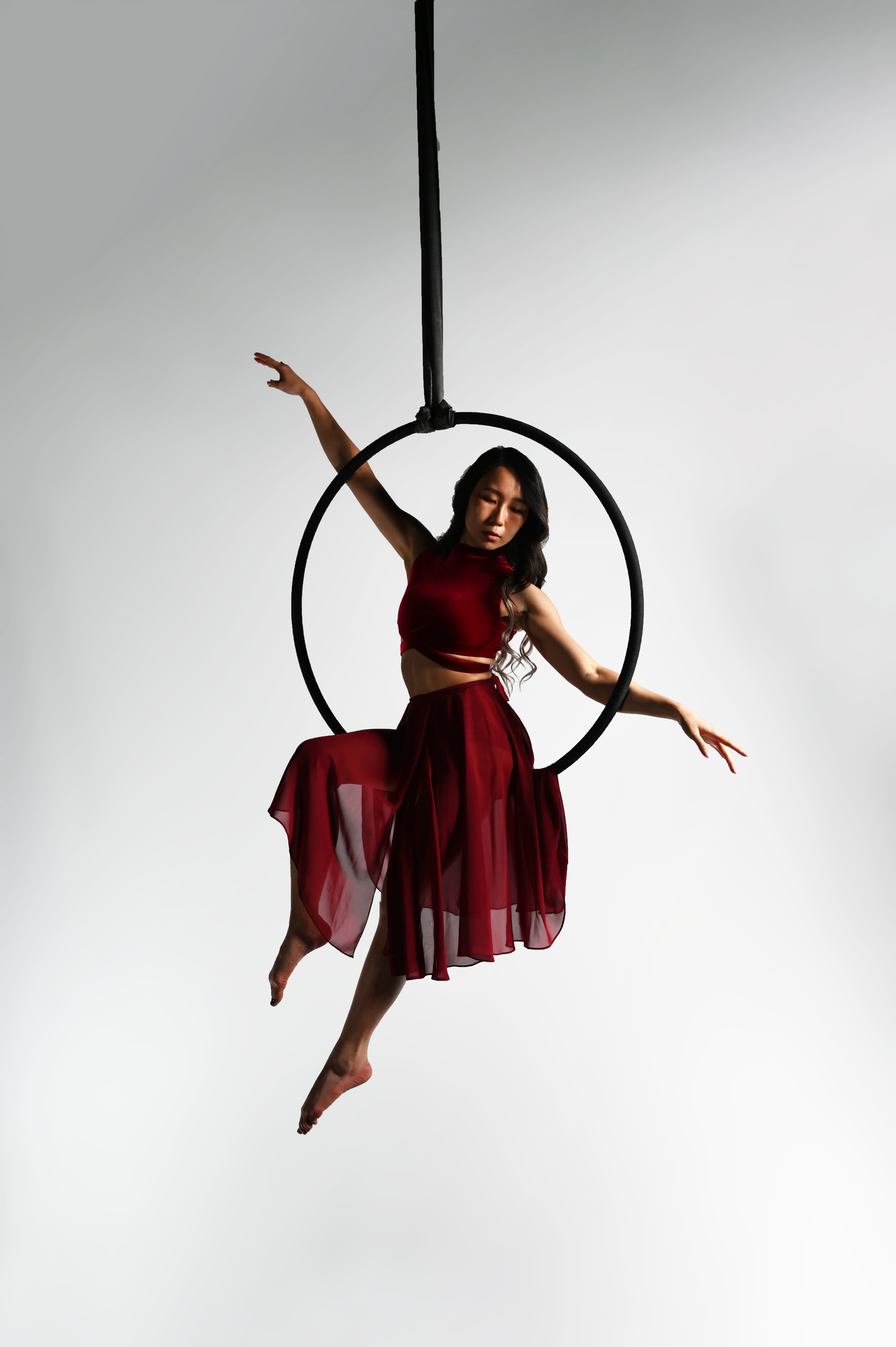 A woman performing aerial hoop on a black hoop suspended from the ceiling, wearing a flowing red dress, against a plain white background.