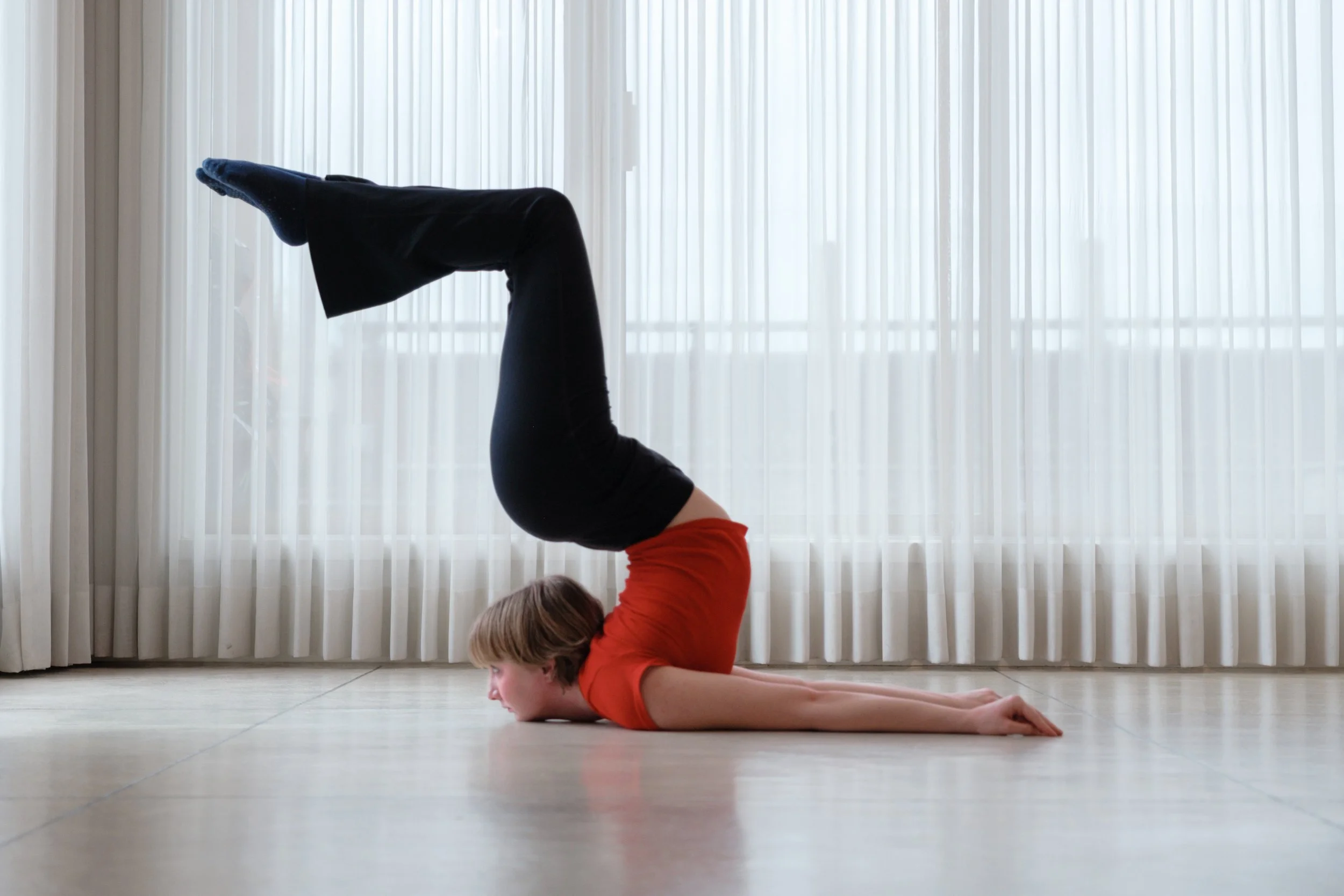 Woman doing a contortion cheststand yoga pose in a room with large windows and sheer curtains.