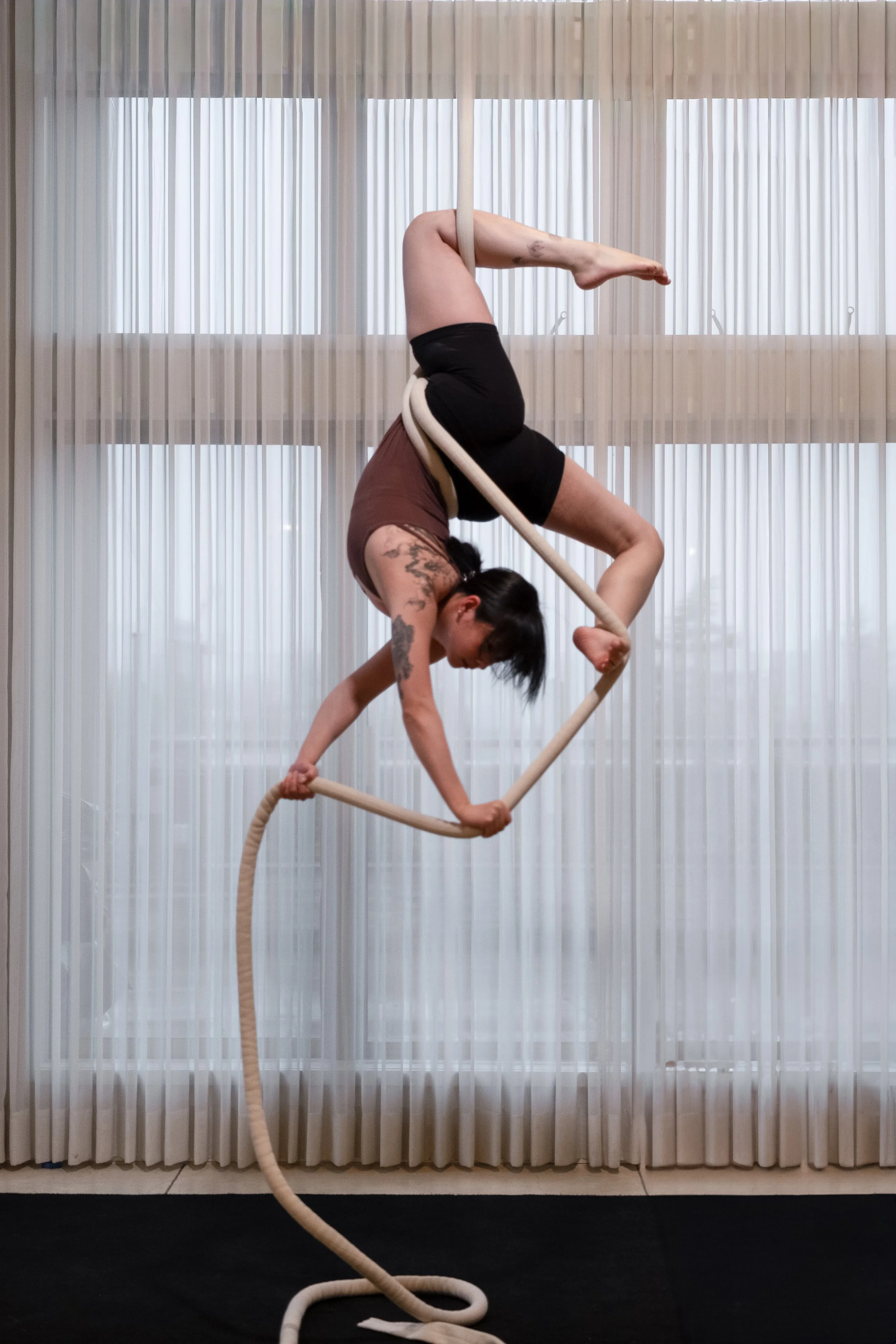 A woman performing aerial rope acrobatics inside near a window with curtains.