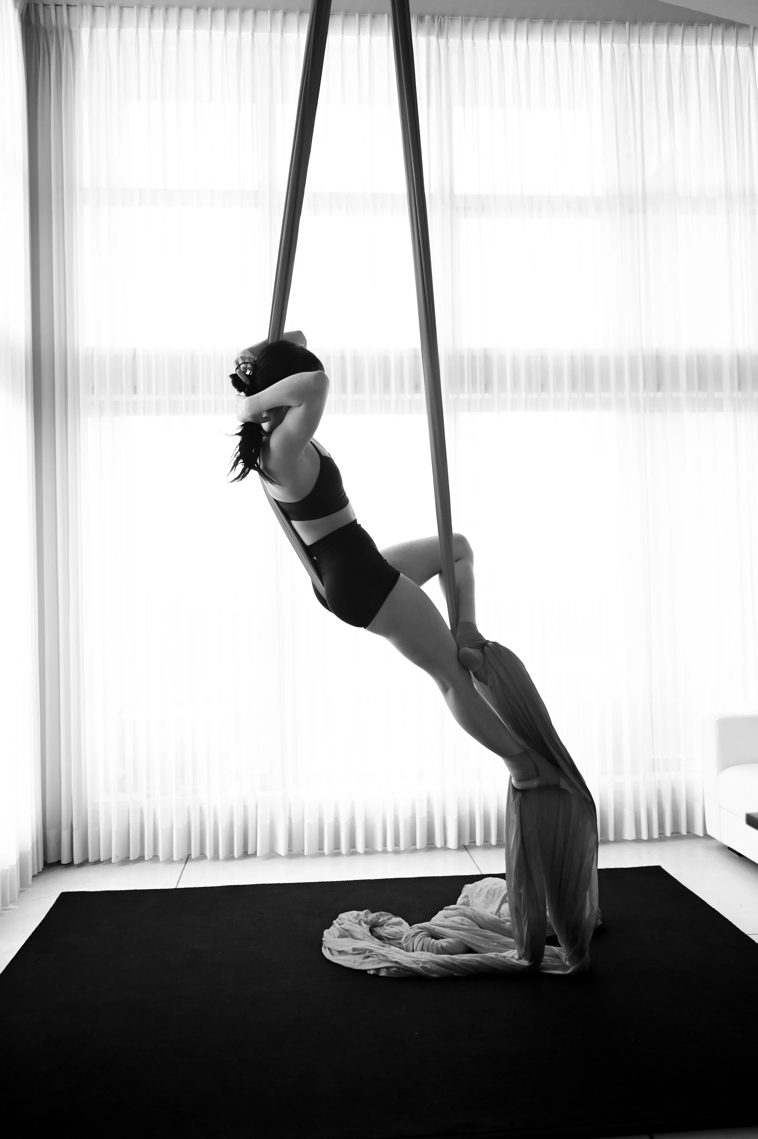 A woman practicing aerial silks yoga indoors with natural light coming through large curtains.