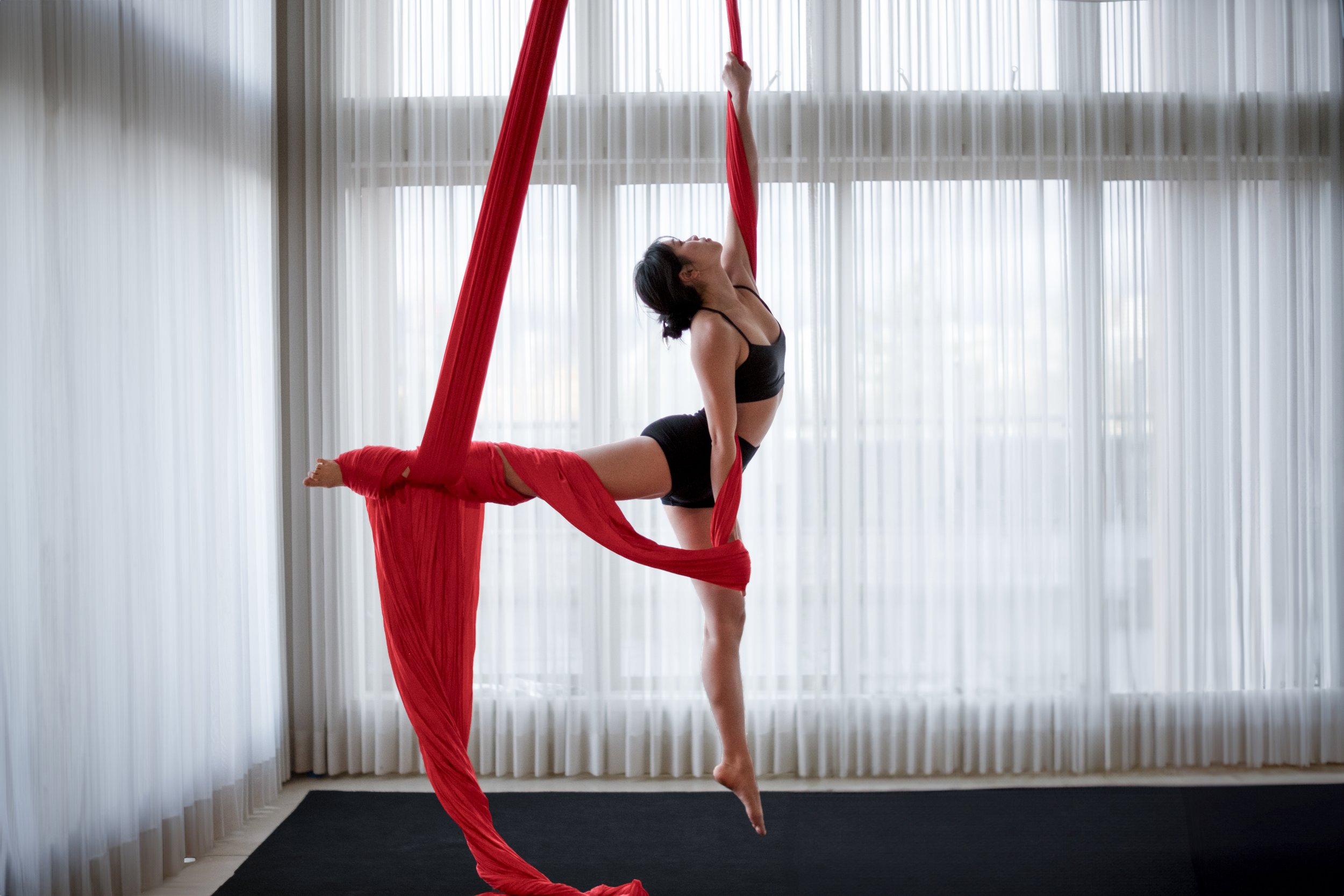 A woman performing an aerial silk yoga pose in a bright room with floor-to-ceiling curtains, hanging from red silk fabric.