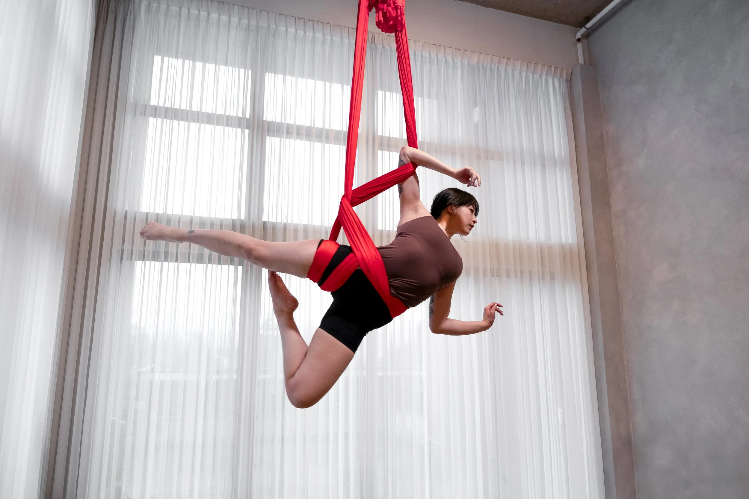 Woman performing aerial hammock yoga pose indoors, hanging from red fabric suspended from ceiling near large window with white curtains.