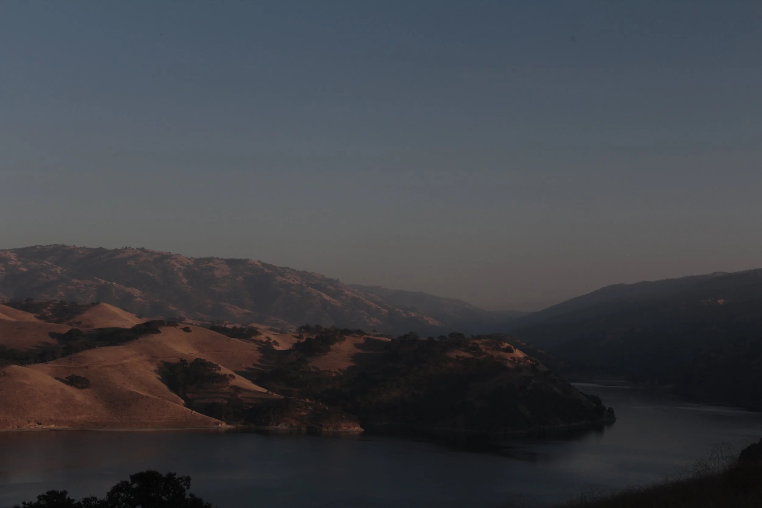 Scenic view of rolling hills and mountains surrounding a river under a clear sky at dawn or dusk.