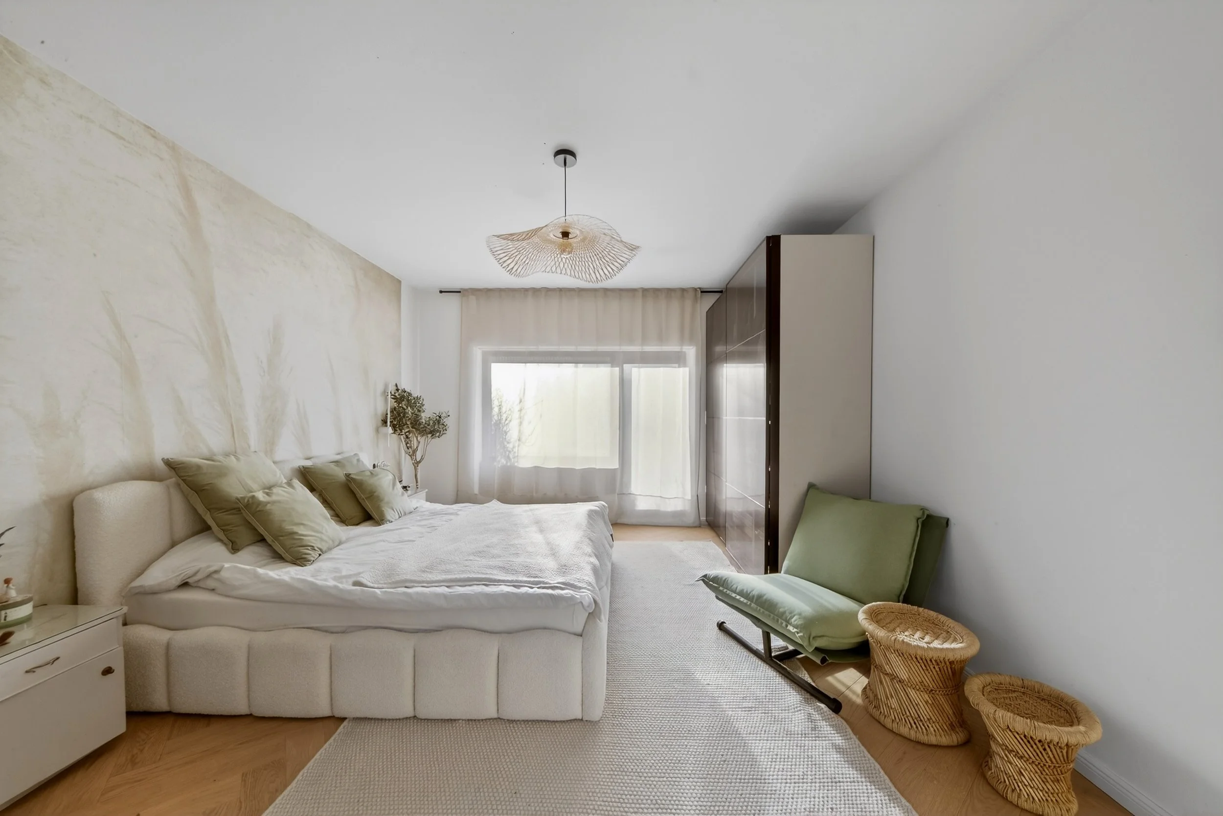 A bedroom with a white bed, green pillows, beige curtains, a green lounge chair, two woven stools, and a small bedside table with a plant, natural light coming through the window.