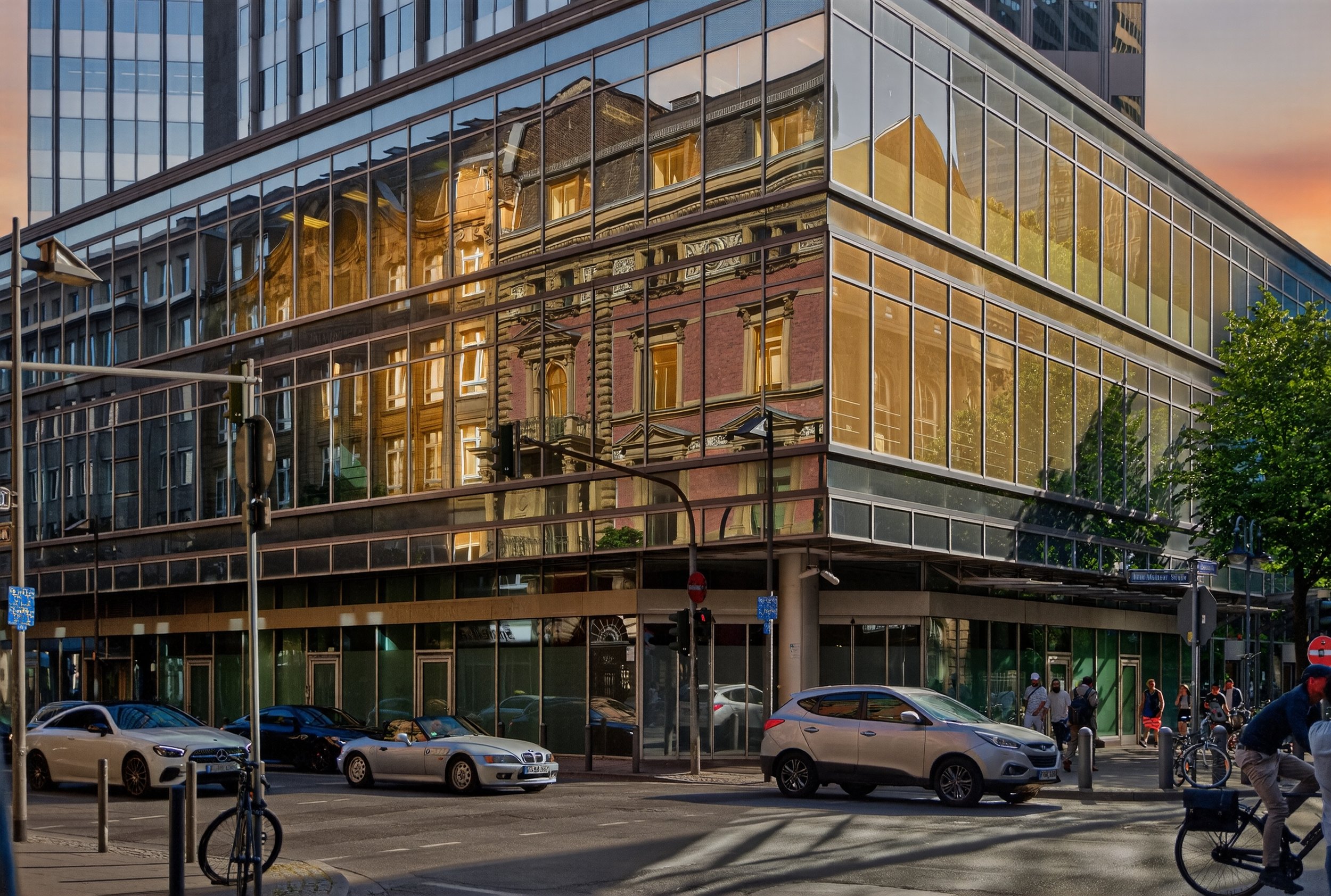 Modern glass building reflecting historic architecture across the street, with cars and pedestrians on city street at sunset.