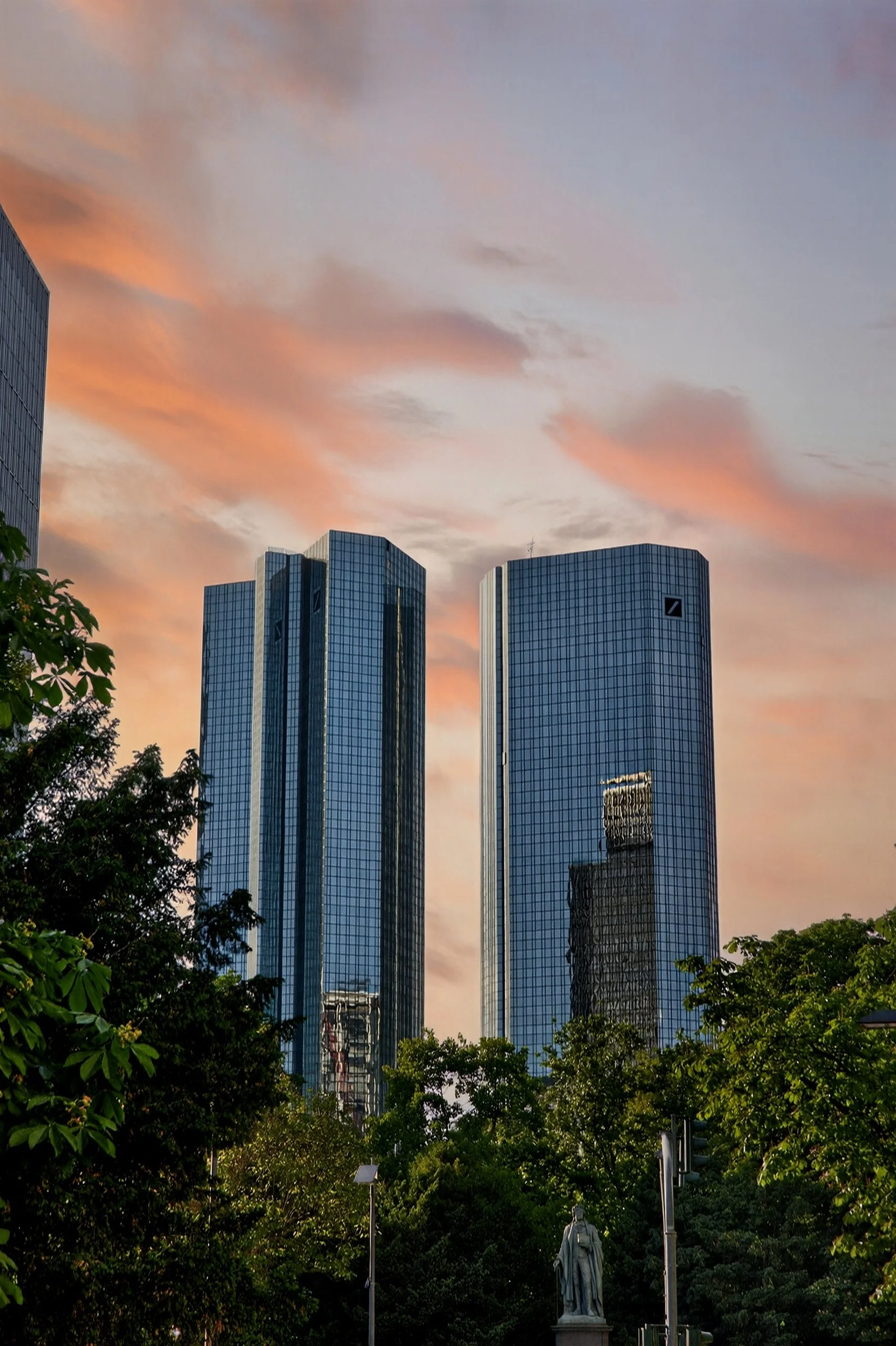 Two modern skyscrapers with reflective glass facades during sunset, surrounded by green trees and a statue in the foreground.