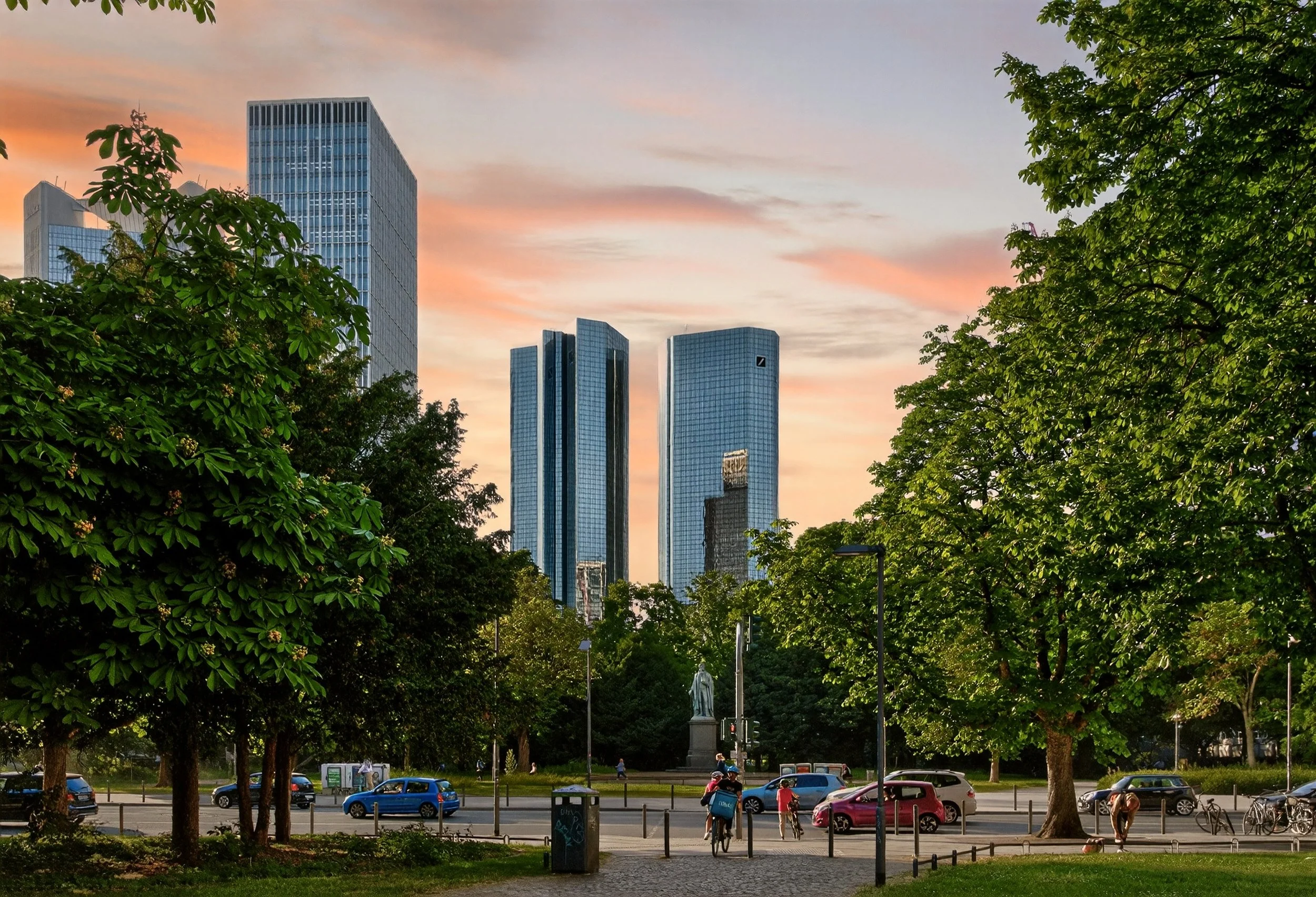 City park with trees, people biking, and cars parked along the street, with modern glass skyscrapers in the background during sunset.