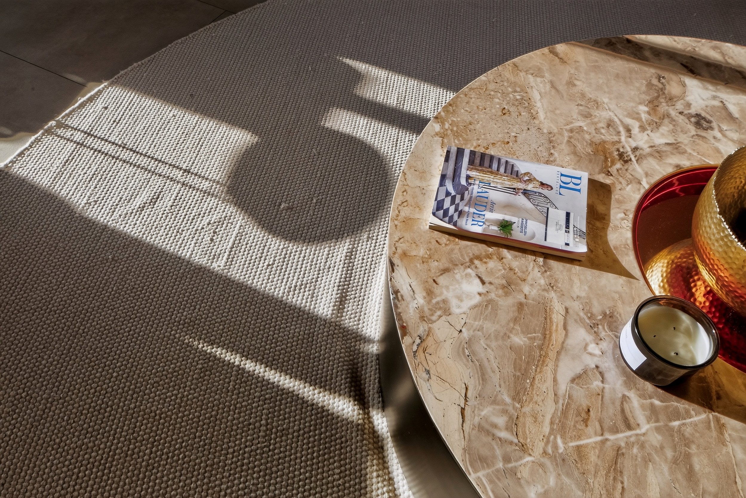 Close-up of a marble coffee table with a magazine, a candle, and a decorative vase. Sunlight creates shadows on a textured rug nearby.Number one real estate photography agency in Rwanda, real estate photographer in Germany.