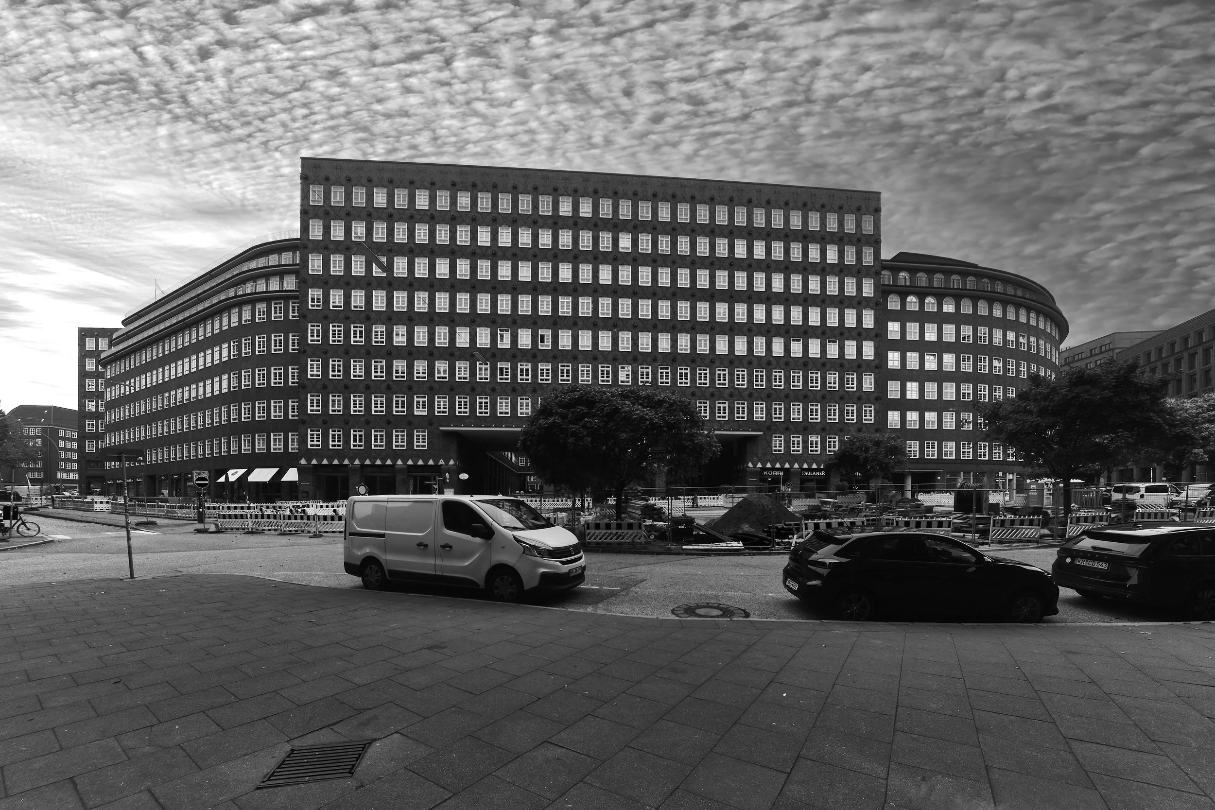Black and white photo of a large multi-story brick building with many windows, situated on a city street with some parked cars and trees in front.