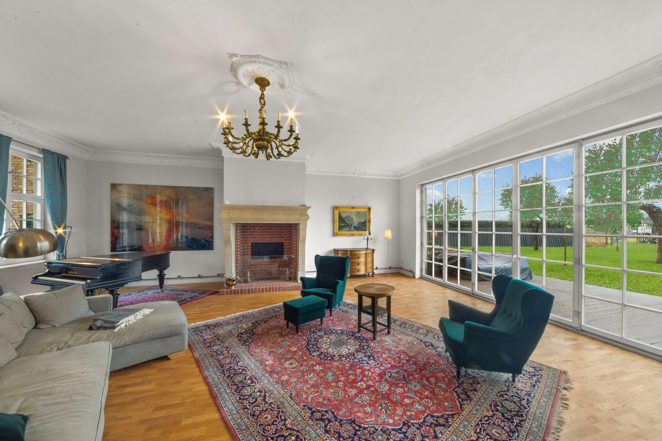 A spacious living room with large windows showing a green yard outside. The room has a white ceiling with crown molding, a chandelier, a brick fireplace, and light-colored walls. It features a beige sofa, two teal armchairs, a colorful Persian rug, a