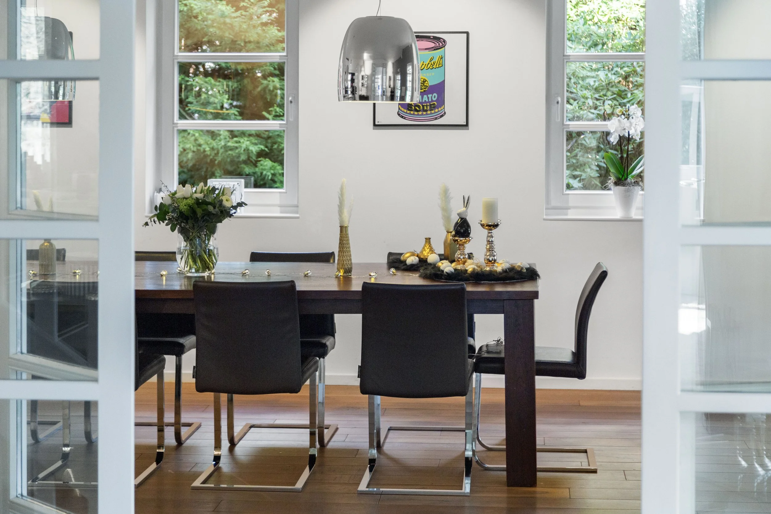 Dining room with a wooden table, six black chairs, floral centerpiece, and festive decorations, viewed through open glass double doors with windows showing green trees outside.