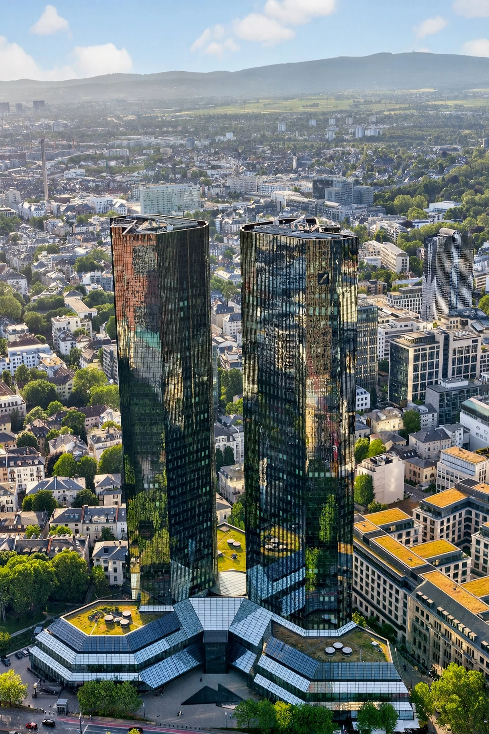 Two tall, reflective glass skyscrapers in an urban city from an aerial view, with a sprawling cityscape and hills in the background.