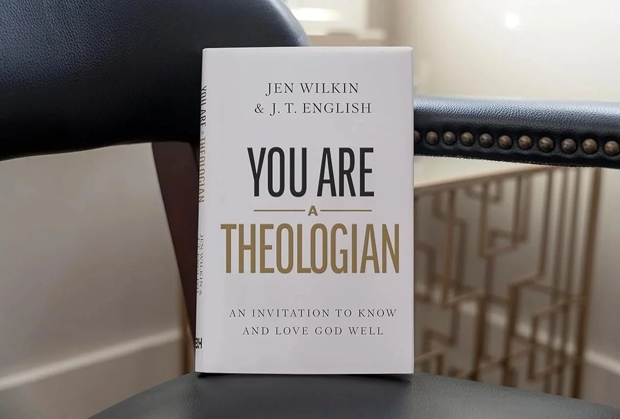 Close-up of a book titled "You Are a Theologian" by Jen Wilkin and J. T. English, placed on a black chair with a metal decorative side panel in the background.