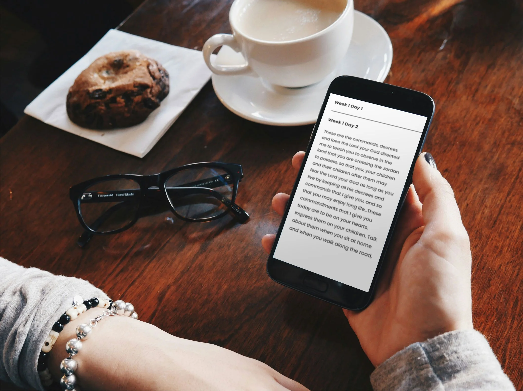 A person is holding a smartphone with a religious text displayed on the screen. On a wooden table in front of them, there are black glasses, a chocolate chip cookie on a paper napkin, and a cup of coffee or tea in a white cup and saucer.