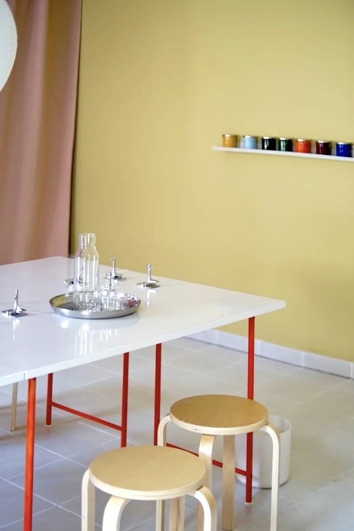 Ein weißer Tisch mit zwei runden Holzscheiben als Sitzflächen, Gläser und eine silberne Schale auf dem Tisch, gelbe Wand mit einer weißen Wandregal mit bunten Farbgläsern, rote Tischbeine, pinke Vorhänge im Hintergrund.