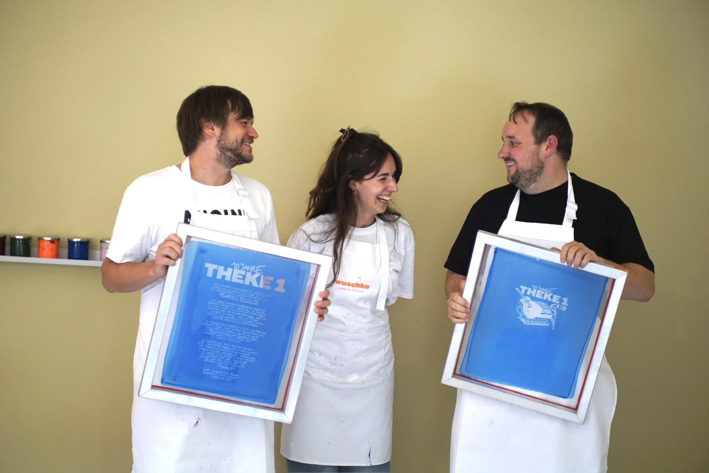 Drei Menschen lachen und halten große blaue Siebdruckrahmen mit dem BAndlogo von "Theke 1", während sie auf eine gelbe Wand im Hintergrund schauen.
