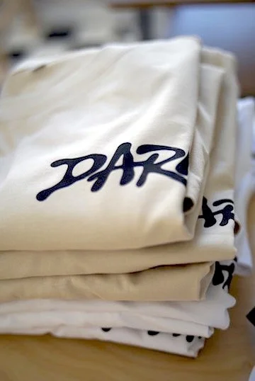 Mehrere weiße Textilien mit dem schwarzen Logo DARI gestapelt auf einem Tisch.
