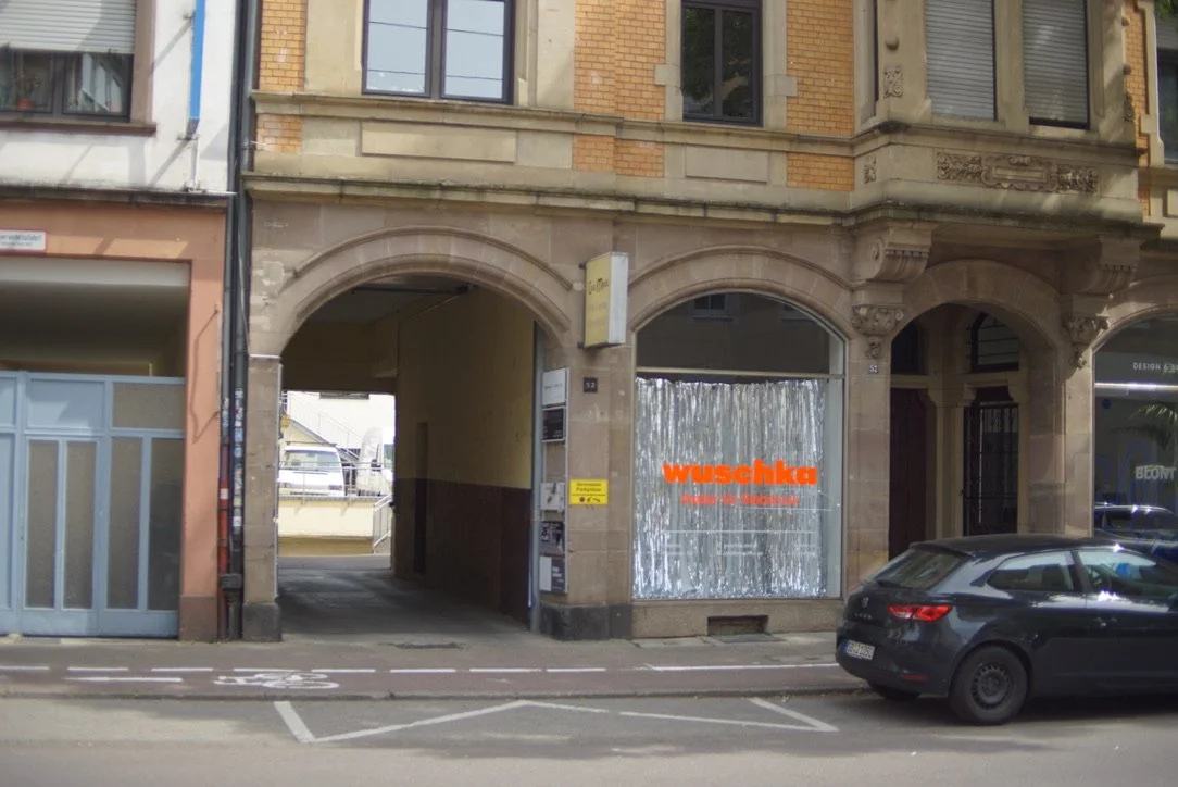 Mainzerstraße saarbrücken. Ein Altbaugebäude mit einem Durchgang in der Mitte, links befindet sich eine blaue Tür, rechts eine große Schaufensterfront mit einem aufgebrachten silbernen Tuch und einem roten wuschka Schriftzug.