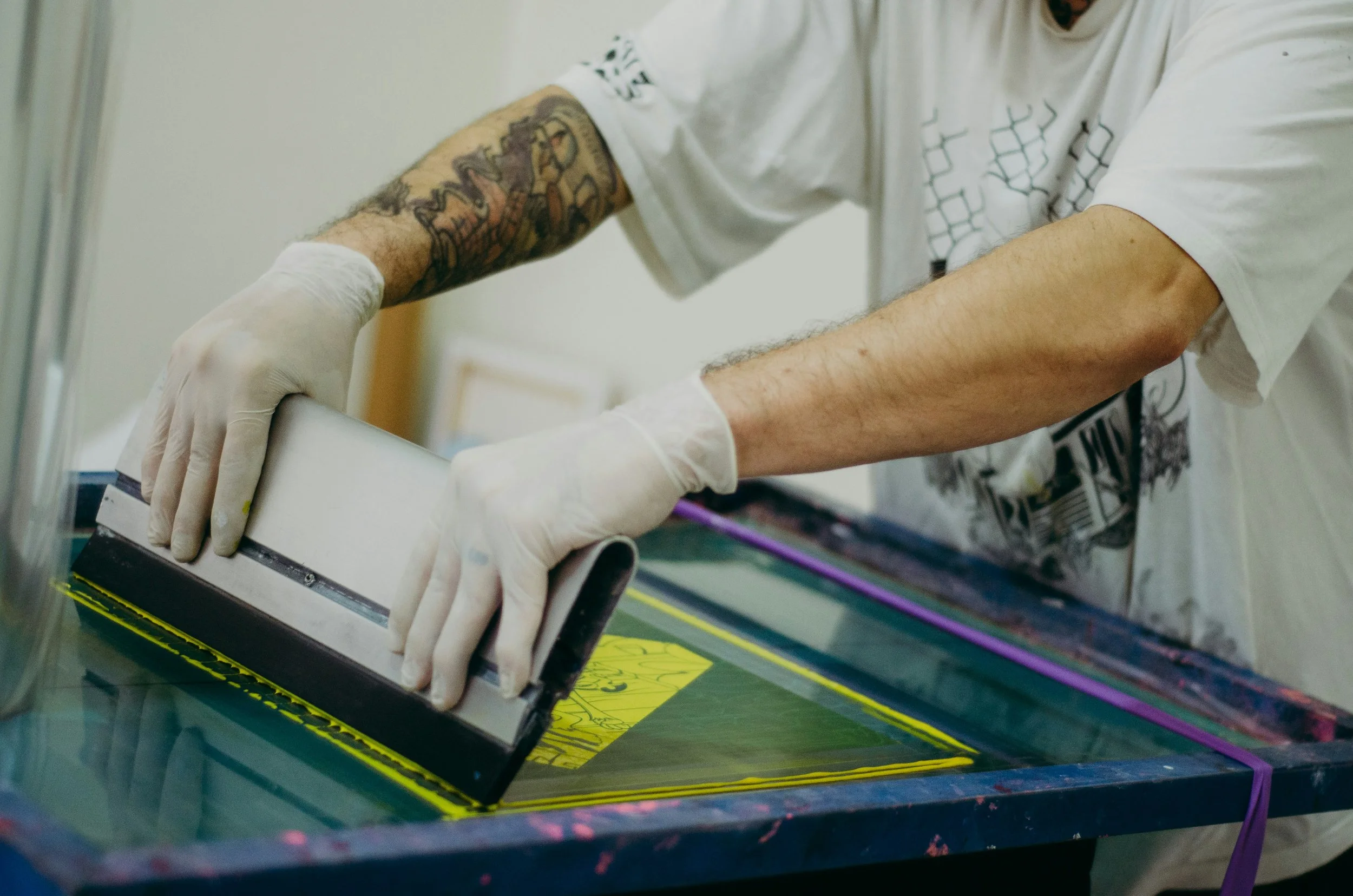 Beim Siebdruck entstehen durch Handarbeit besonders intensive Farben und individuelle Ergebnisse. Person bei der Tätowierung, der eine Schablone auf die Haut überträgt, trägt Handschuhe, hat Tattoos am Arm und trägt ein T-Shirt.